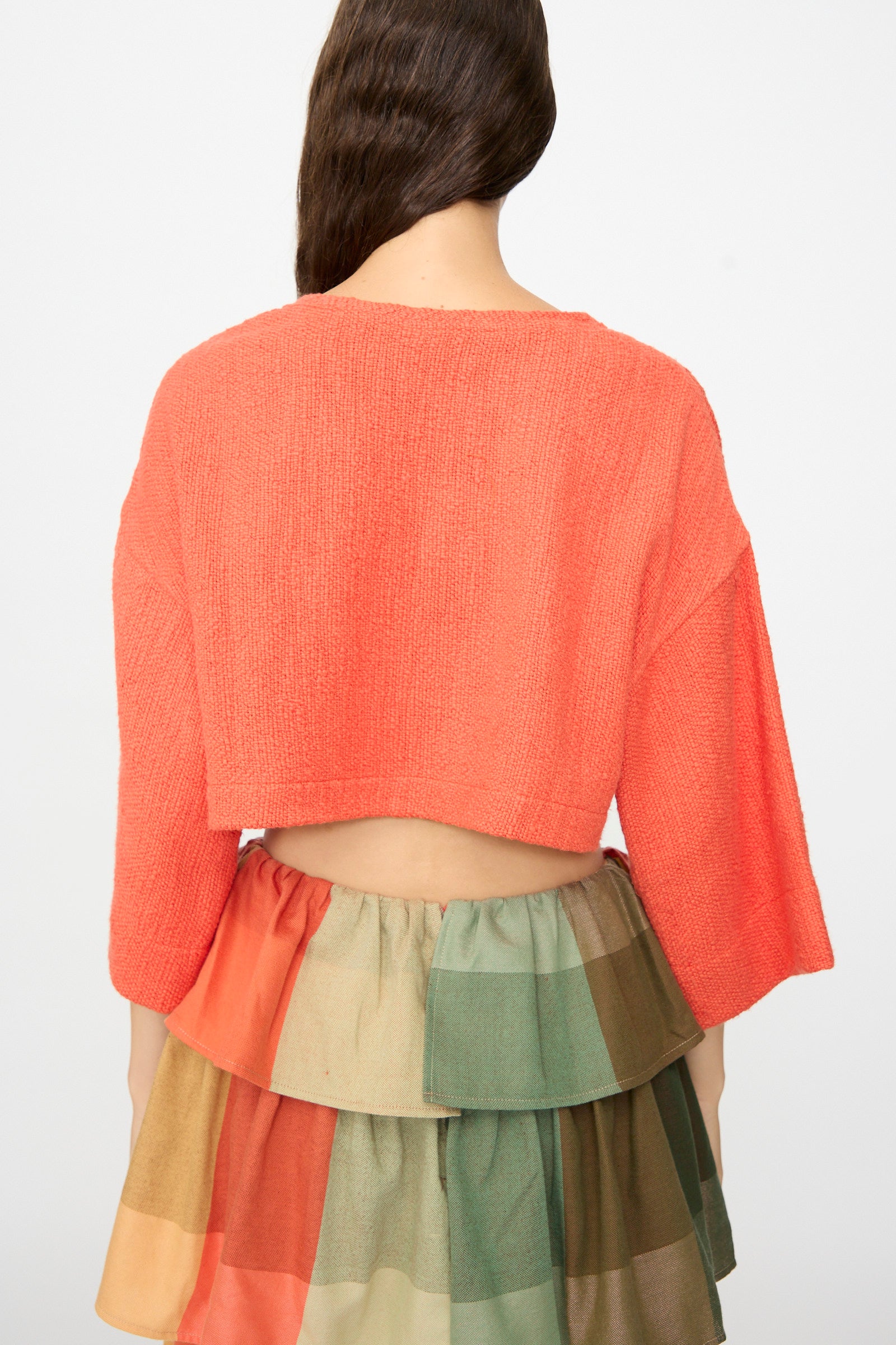 A person is facing away, wearing The Cropped T-Shirt in Burnt Orange Flamme by Marrakshi Life, styled with wide sleeves and paired with a multicolored, tiered checkered skirt crafted from handwoven cotton. The backdrop is plain and light-colored.