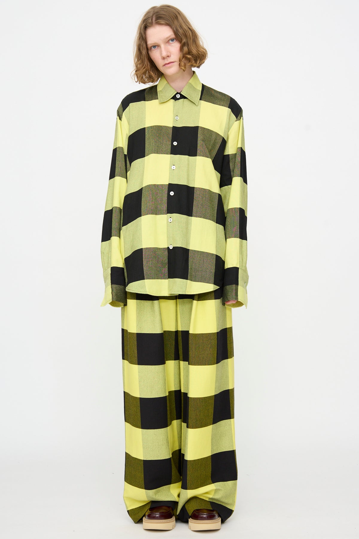 A person stands against a white background, wearing Marrakshi Life’s The French Cuff Shirt in Plaid Black and Sunny—an oversized cotton shirt with long sleeves—styled with matching wide-leg pants and brown platform shoes.