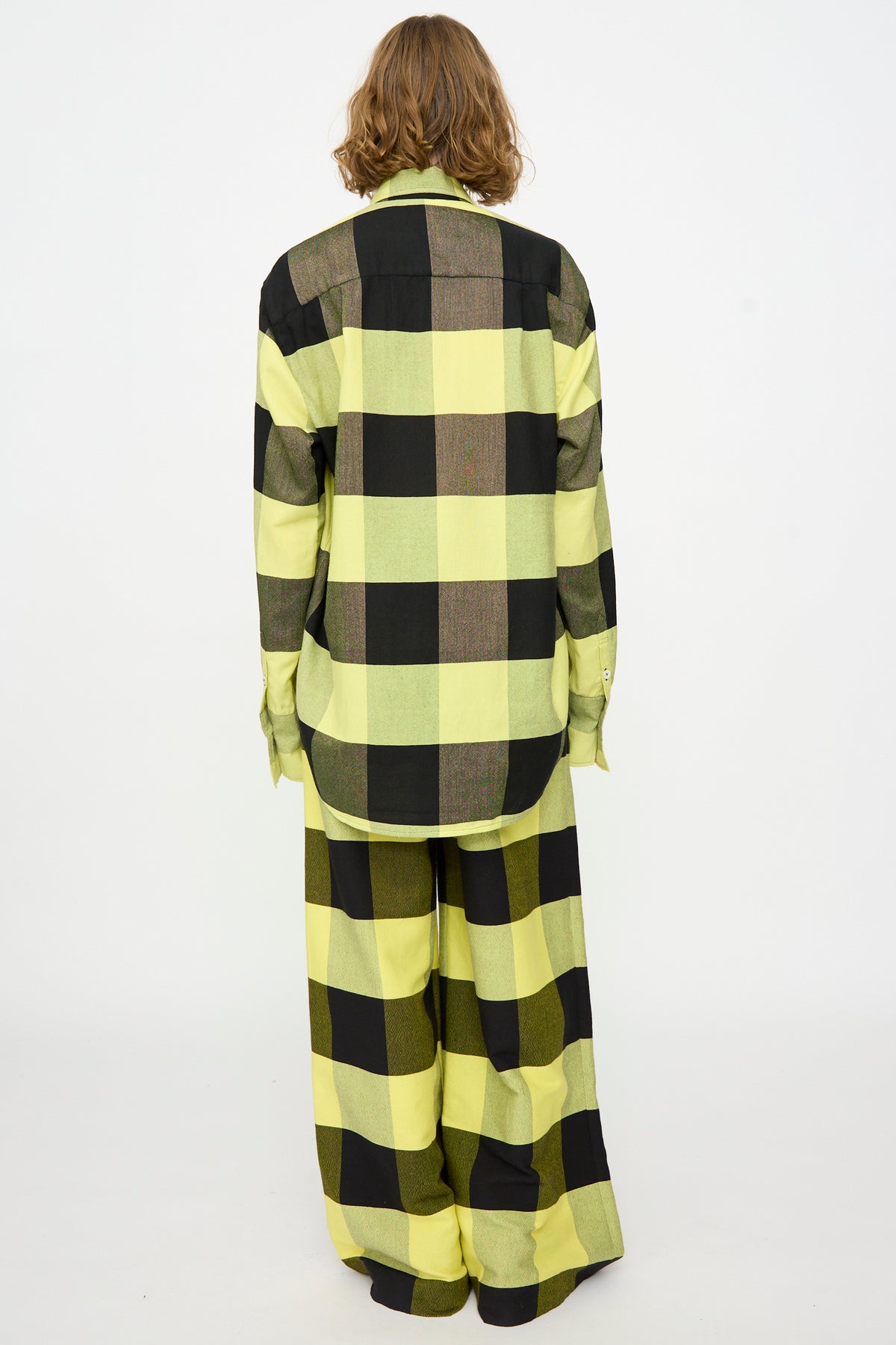 A person stands with their back to the camera, wearing Marrakshi Life’s The French Cuff Shirt in Plaid Black and Sunny, paired with wide-leg pants, against a plain white background.