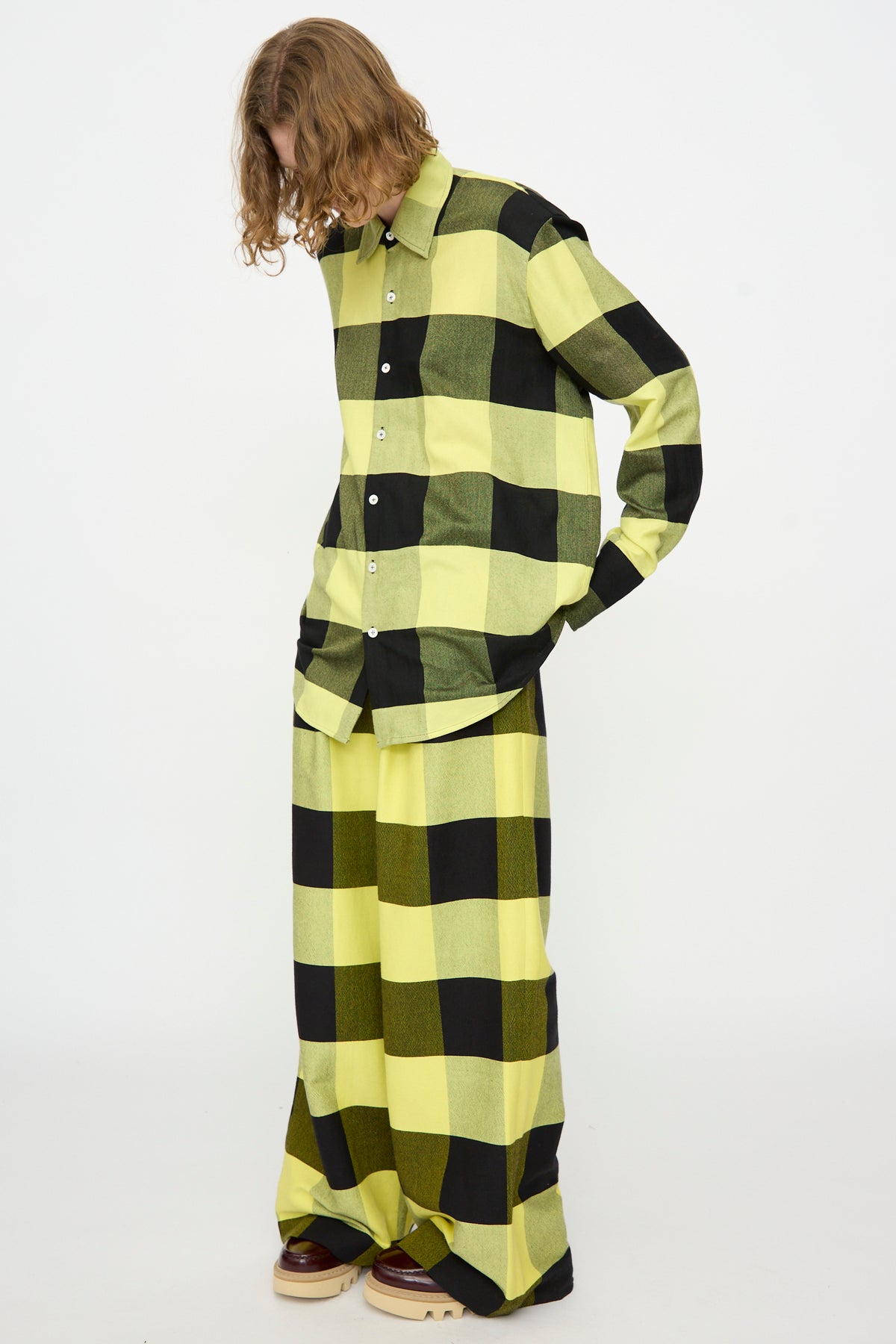 Wearing Marrakshi Life’s The French Cuff Shirt in Plaid Black and Sunny, this person pairs the yellow and black cotton top with matching wide-legged pants, looking down against a plain white background.