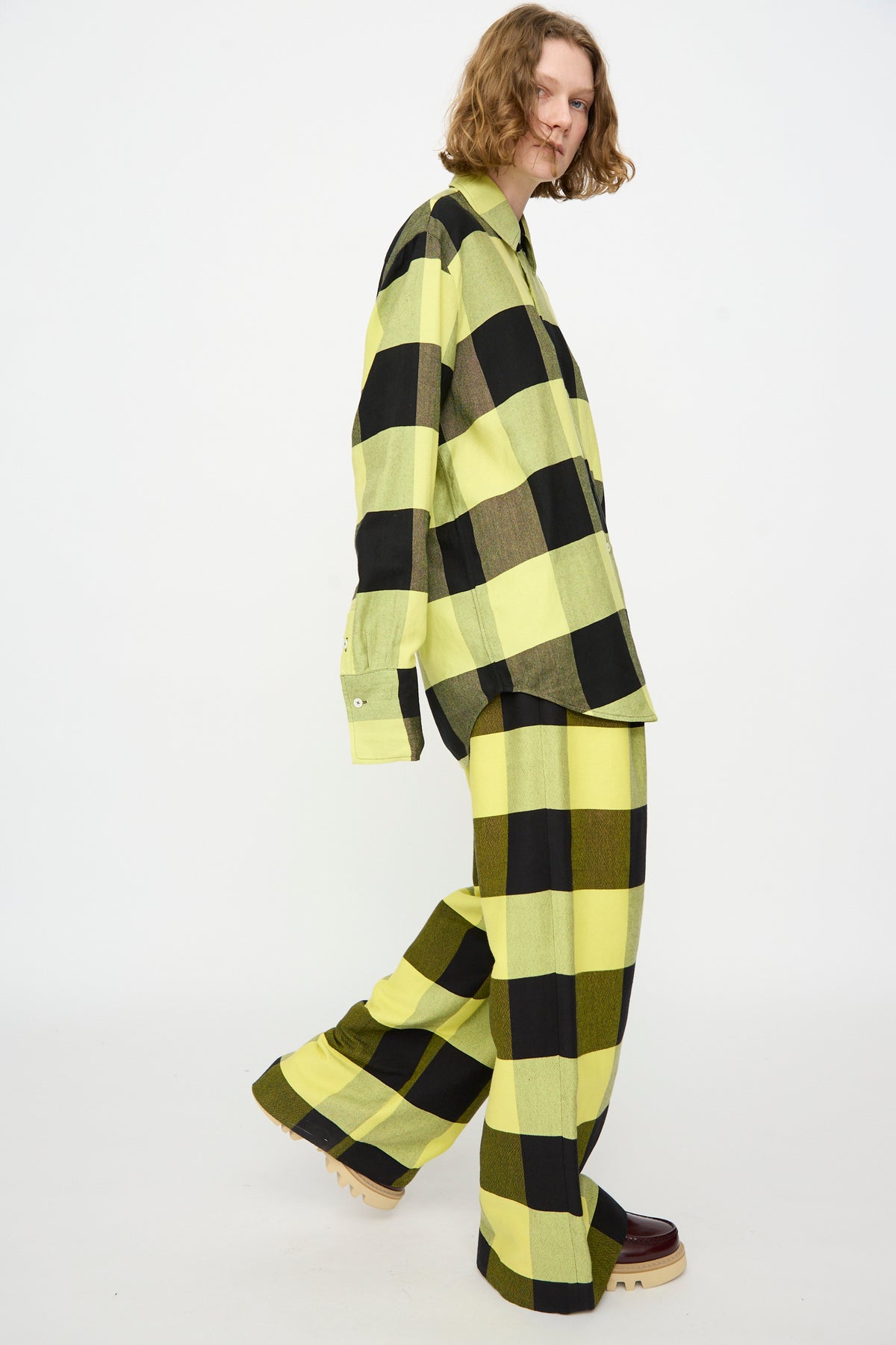 A person wearing Marrakshi Life’s The French Cuff Shirt in Plaid Black and Sunny—an oversized long sleeve cotton shirt in yellow and black plaid—paired with matching wide-leg pants and beige platform shoes, standing against a plain white background.