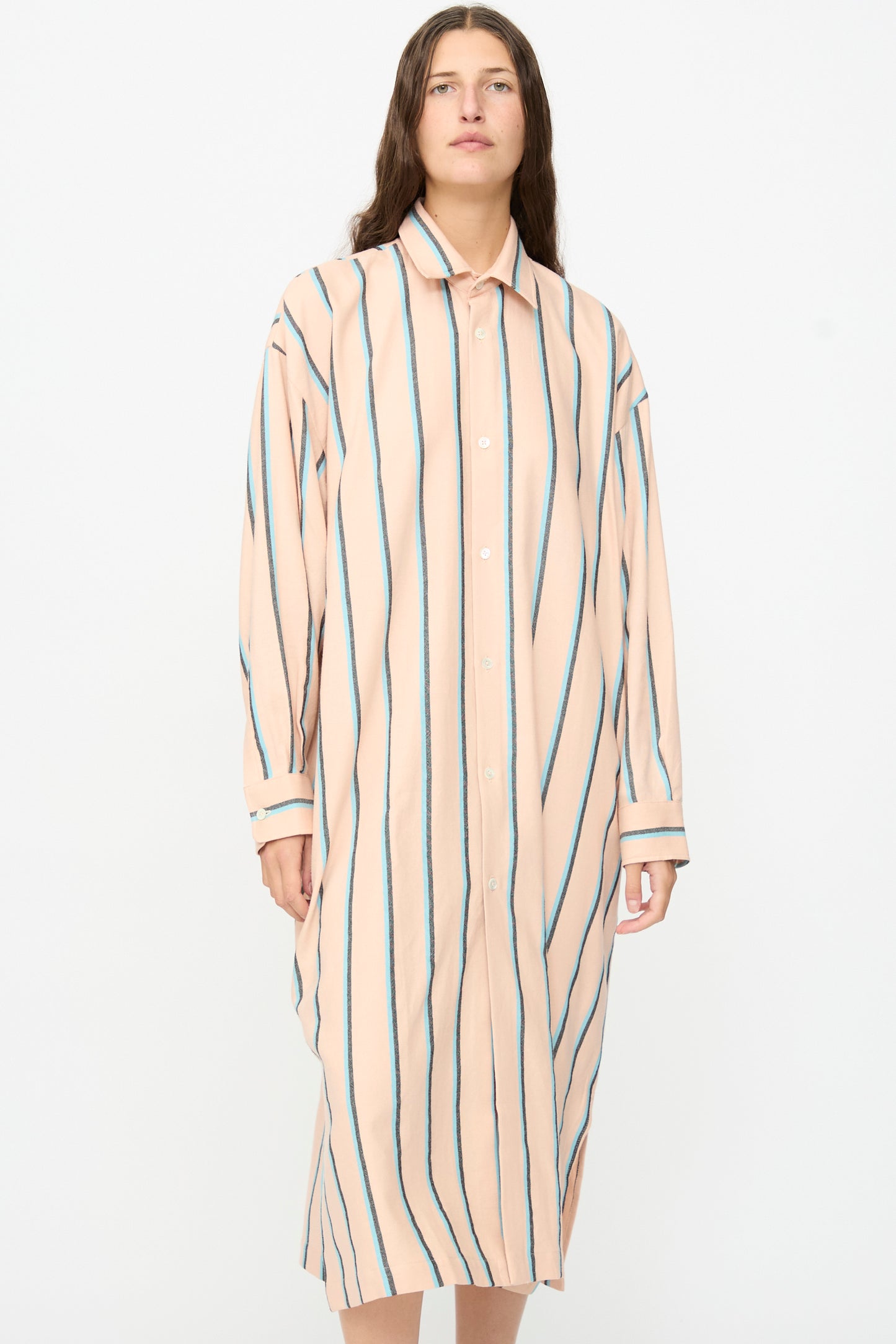 A person stands against a plain background, wearing Marrakshi Life’s The Long Shirt in Stripe—an oversized, peach-colored button-down dress featuring vertical green and blue stripes.