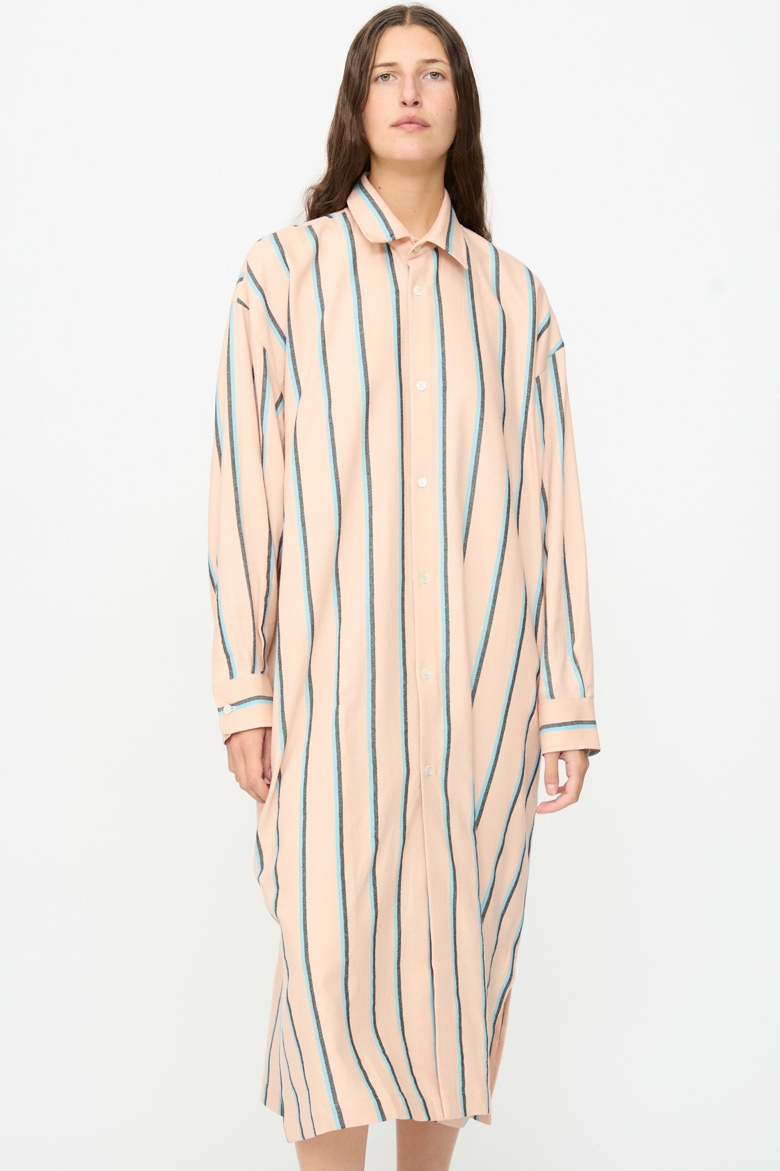 A person stands against a plain background, wearing Marrakshi Life’s The Long Shirt in Stripe—an oversized, peach-colored button-down dress featuring vertical green and blue stripes.