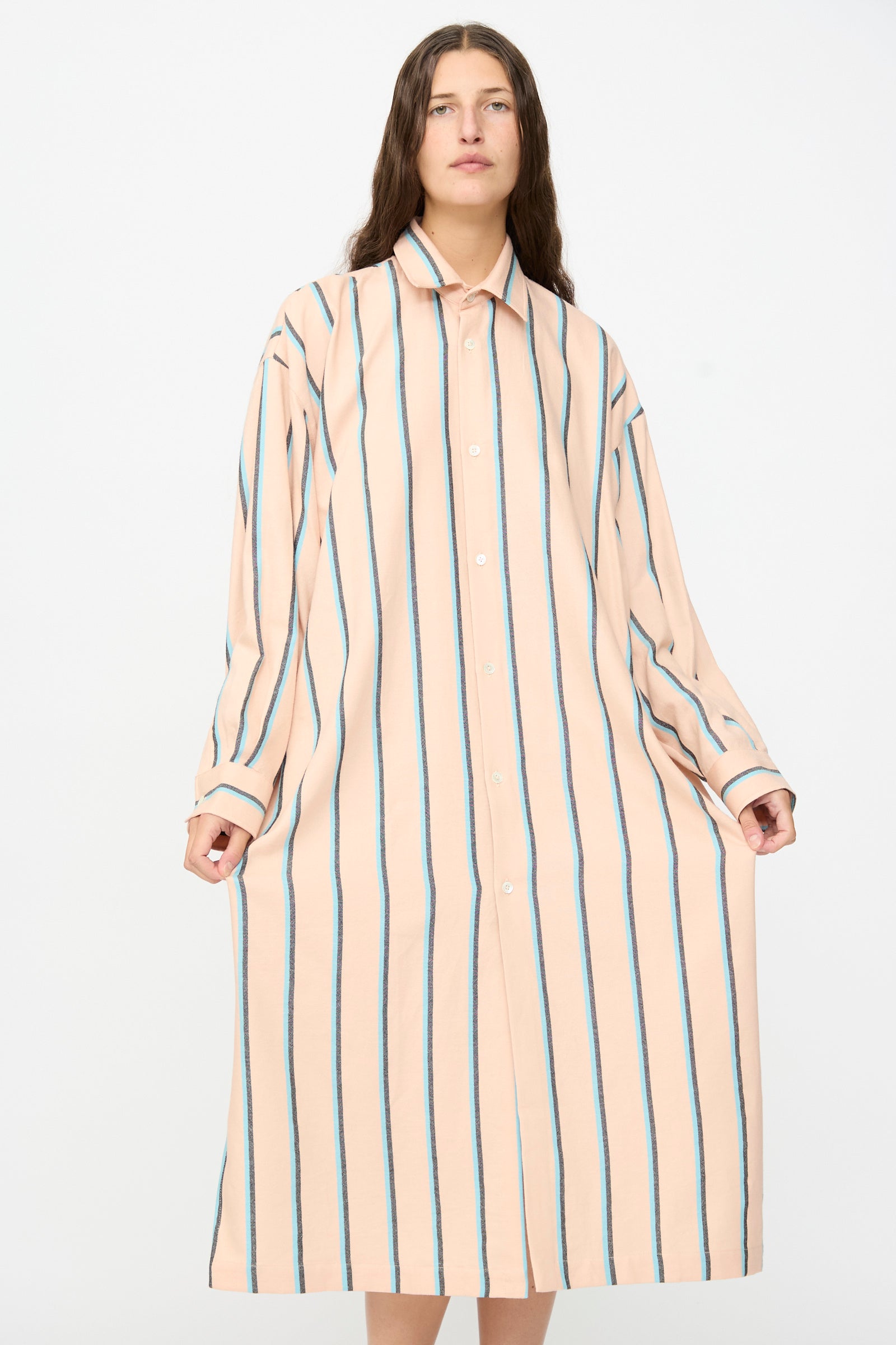 A person wearing The Long Shirt in Stripe by Marrakshi Life—an oversized, long-sleeve shirt dress crafted from handwoven cotton in Morocco and featuring vertical blue and black stripes on a peach background—stands against a plain white backdrop.