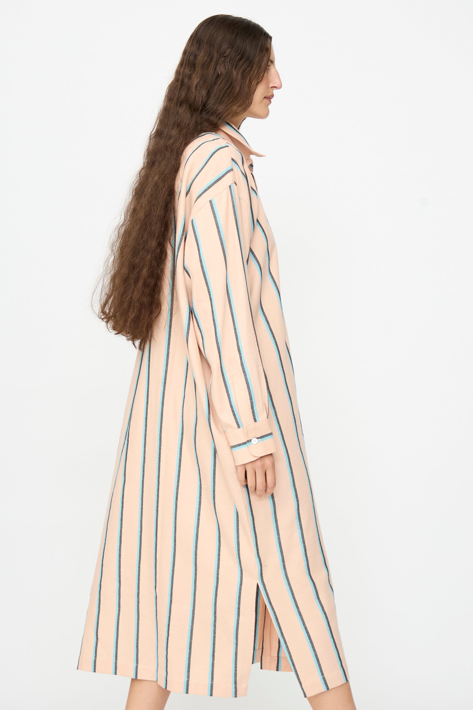 A person wearing Marrakshi Life’s The Long Shirt in Stripe—a long-sleeved, knee-length peach dress with vertical blue stripes reminiscent of an oversized shirt—stands in profile against a plain white background.