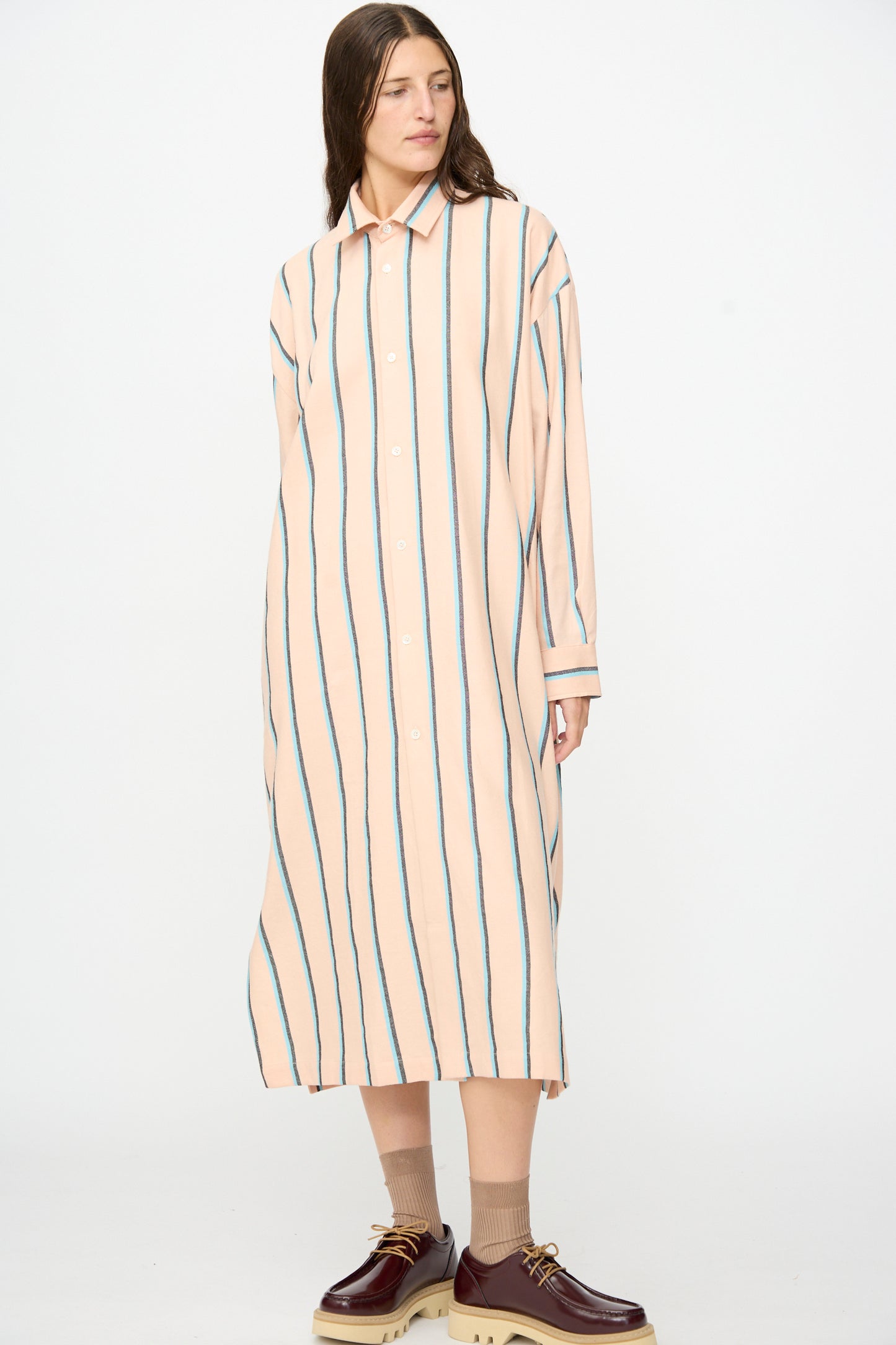 Wearing Marrakshi Life’s The Long Shirt in Stripe—a loose, long, peach-hued dress accented with vertical blue stripes—paired with an oversized long sleeve shirt handmade in Morocco. The outfit is finished with beige socks and burgundy platform shoes, all set against a plain white background.