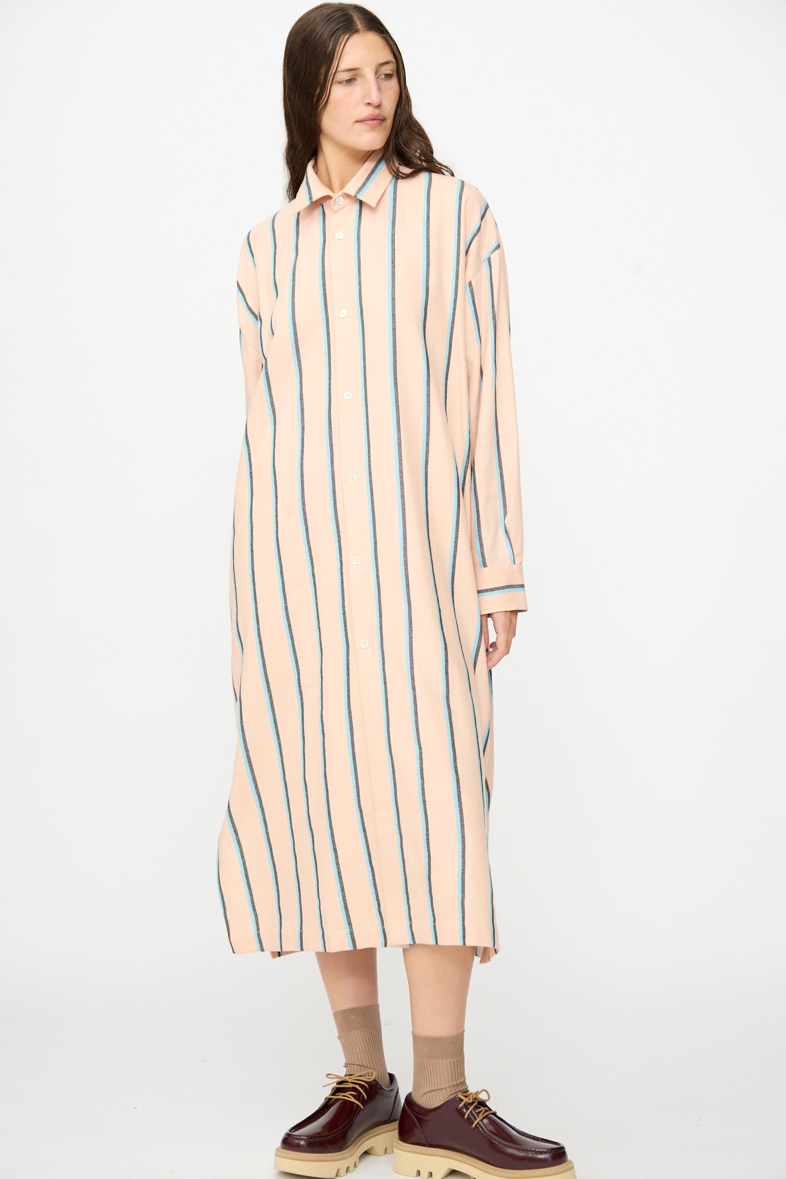 Wearing Marrakshi Life’s The Long Shirt in Stripe—a loose, long, peach-hued dress accented with vertical blue stripes—paired with an oversized long sleeve shirt handmade in Morocco. The outfit is finished with beige socks and burgundy platform shoes, all set against a plain white background.