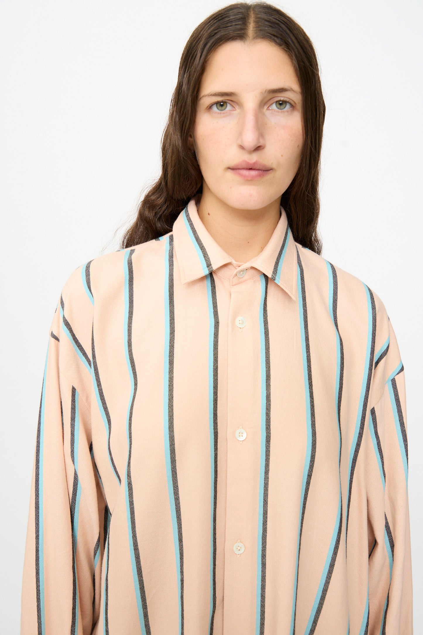 A person with long brown hair wears Marrakshi Life’s The Long Shirt in Stripe—an oversized beige long-sleeve shirt with vertical blue and black stripes—standing against a plain white background.
