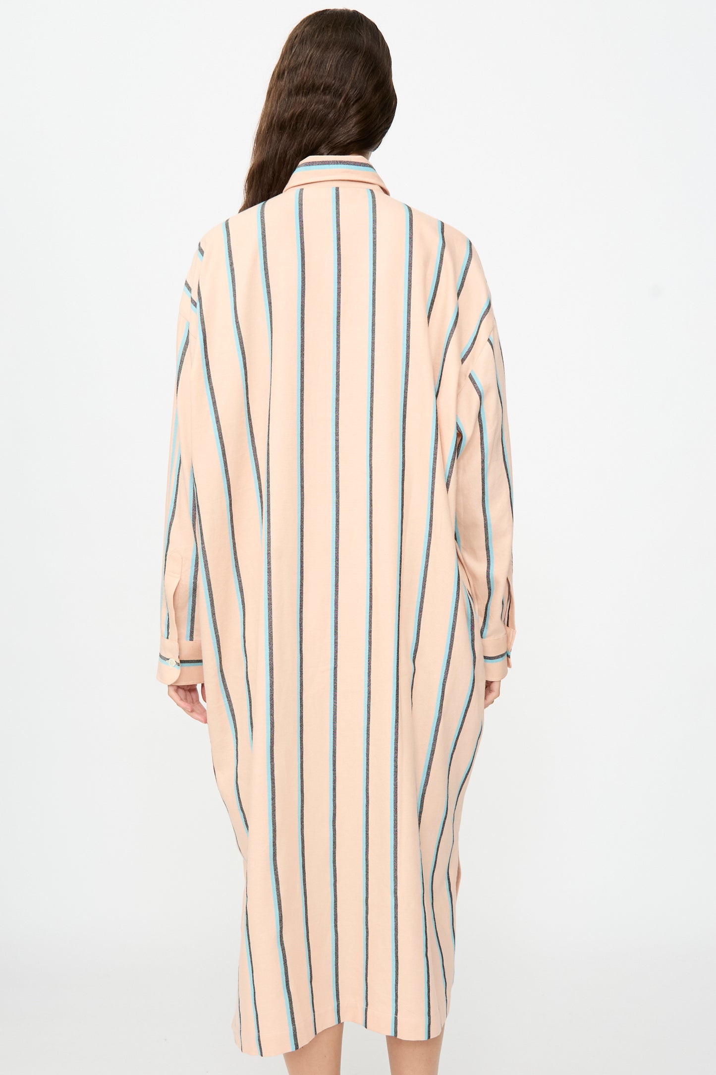 A person with long brown hair is seen from the back against a plain light background, wearing Marrakshi Life’s The Long Shirt in Stripe—an oversized, knee-length dress featuring vertical blue and black stripes and long sleeves.