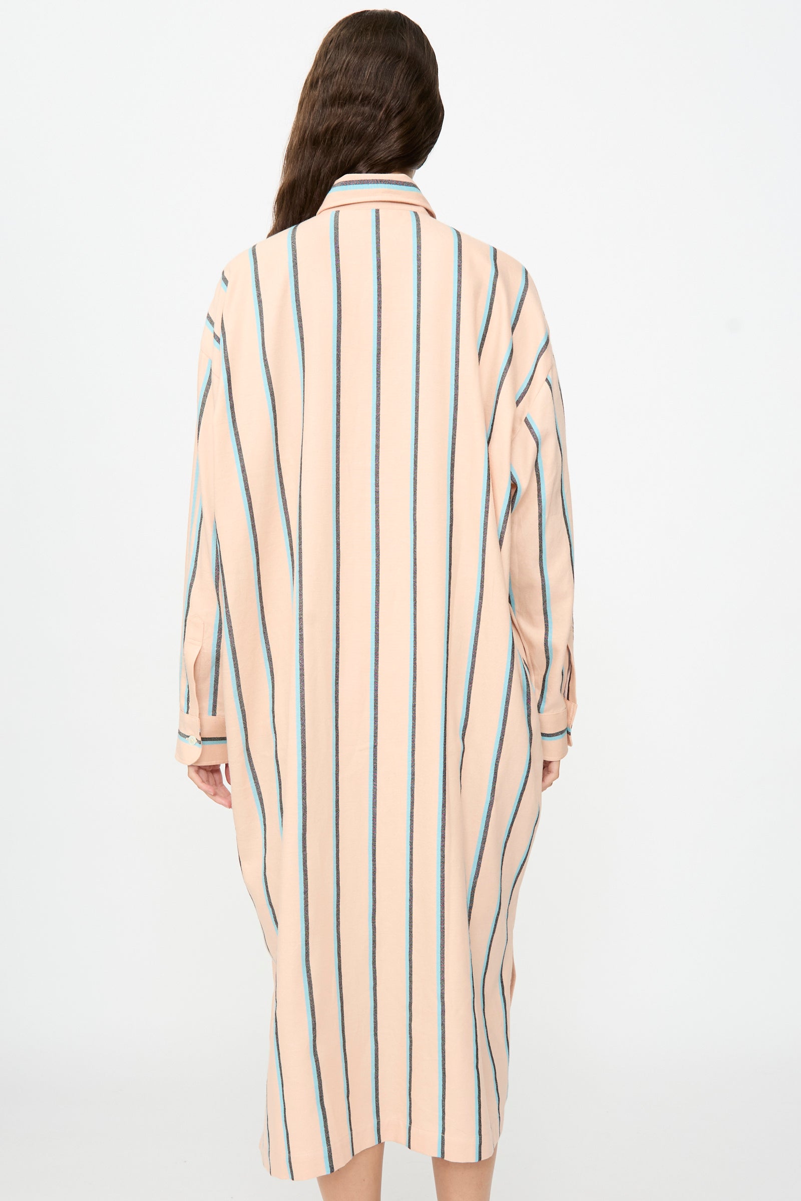 A person with long brown hair is seen from the back against a plain light background, wearing Marrakshi Life’s The Long Shirt in Stripe—an oversized, knee-length dress featuring vertical blue and black stripes and long sleeves.