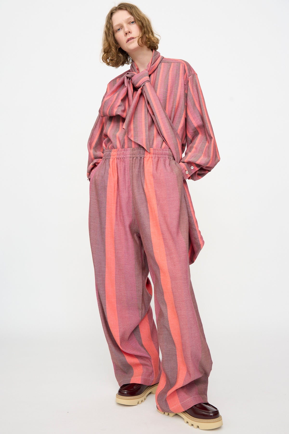 A person is wearing Marrakshi Life’s The Parachute Pant in Stripe Multi—an oversized, wide-leg style paired with a loose, pink and orange striped shirt featuring a large bow, all crafted from handwoven cotton—standing against a plain white background.