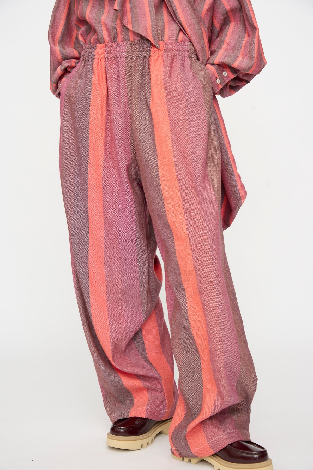 A person wearing Marrakshi Life’s The Parachute Pant in Stripe Multi—an oversized fit, wide-leg pant featuring vertical pink and orange stripes—paired with the matching striped handwoven cotton top and beige platform shoes.