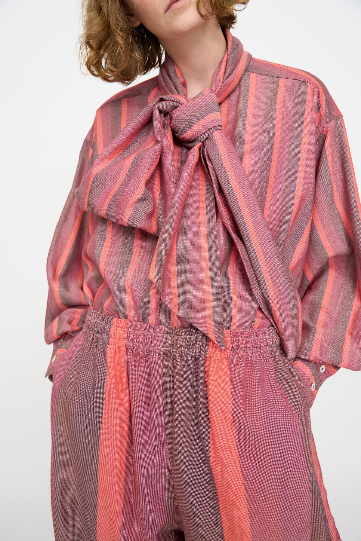 A person wearing Marrakshi Life’s The Parachute Pant in Stripe Multi: a loose-fitting, oversized set with long sleeves and wide-leg pants, covered in vertical pink, purple, and coral stripes and finished with a large bow at the neck.