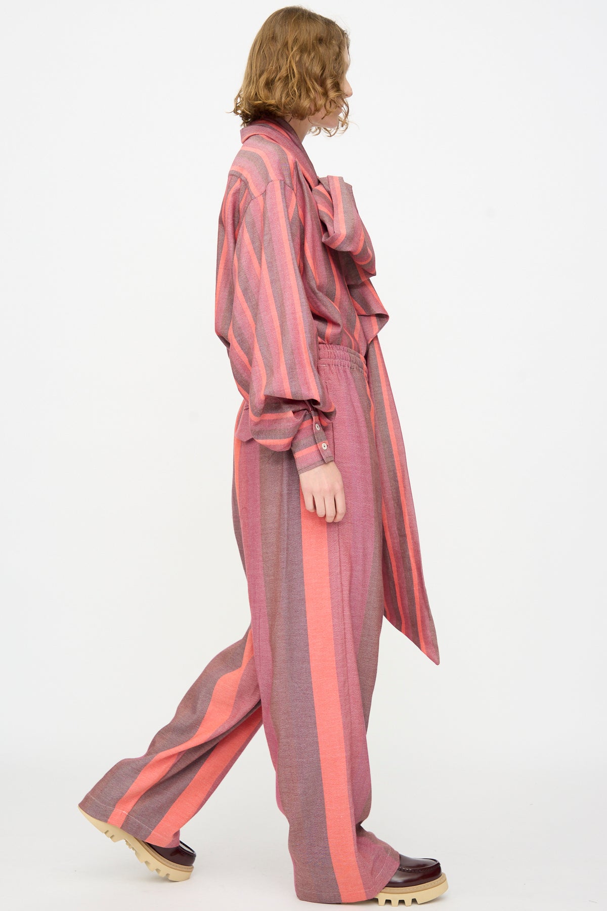 A person stands sideways against a plain white background, wearing Marrakshi Life’s The Parachute Pant in Stripe Multi—an oversized fit, wide-leg pant outfit crafted from soft handwoven cotton in pink and red stripes—paired with platform shoes.