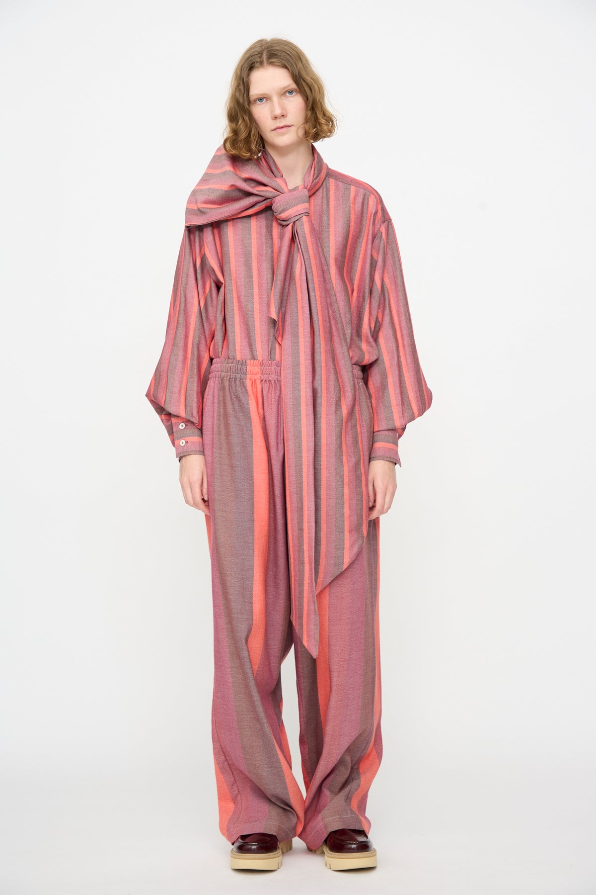 A person stands against a plain white background wearing Marrakshi Life’s The Parachute Pant in Stripe Multi—a loose-fitting, pink and red striped ensemble made from handwoven cotton—styled with an oversized matching scarf tied in a bow.