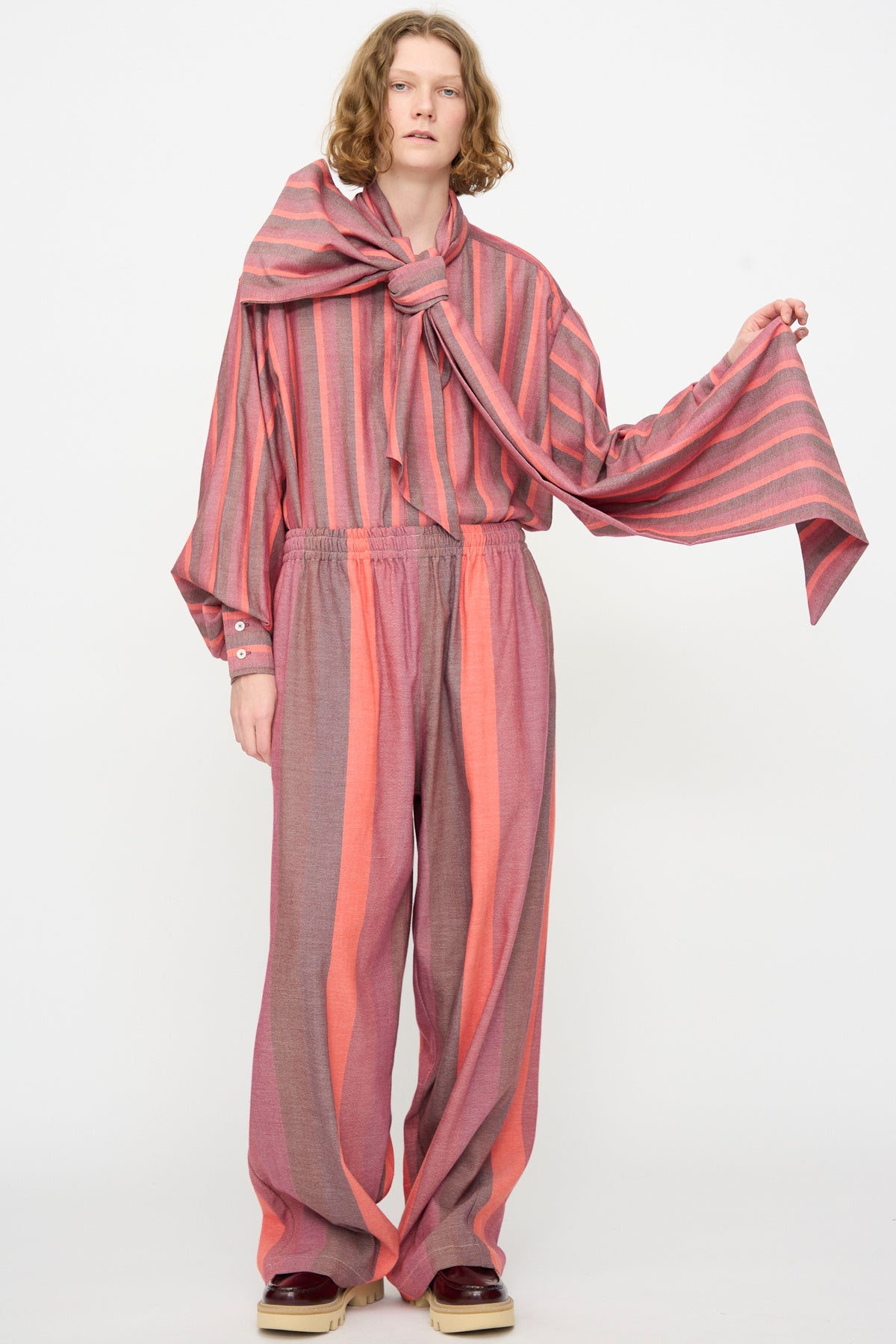 A person standing against a plain background, wearing Marrakshi Life’s The Parachute Pant in Stripe Multi—a loose-fitting, pink and gray striped outfit crafted from handwoven cotton—styled with wide leg pants and a large matching scarf tied at the neck.