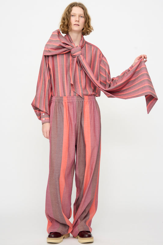 A person standing against a plain background, wearing Marrakshi Life’s The Parachute Pant in Stripe Multi—a loose-fitting, pink and gray striped outfit crafted from handwoven cotton—styled with wide leg pants and a large matching scarf tied at the neck.