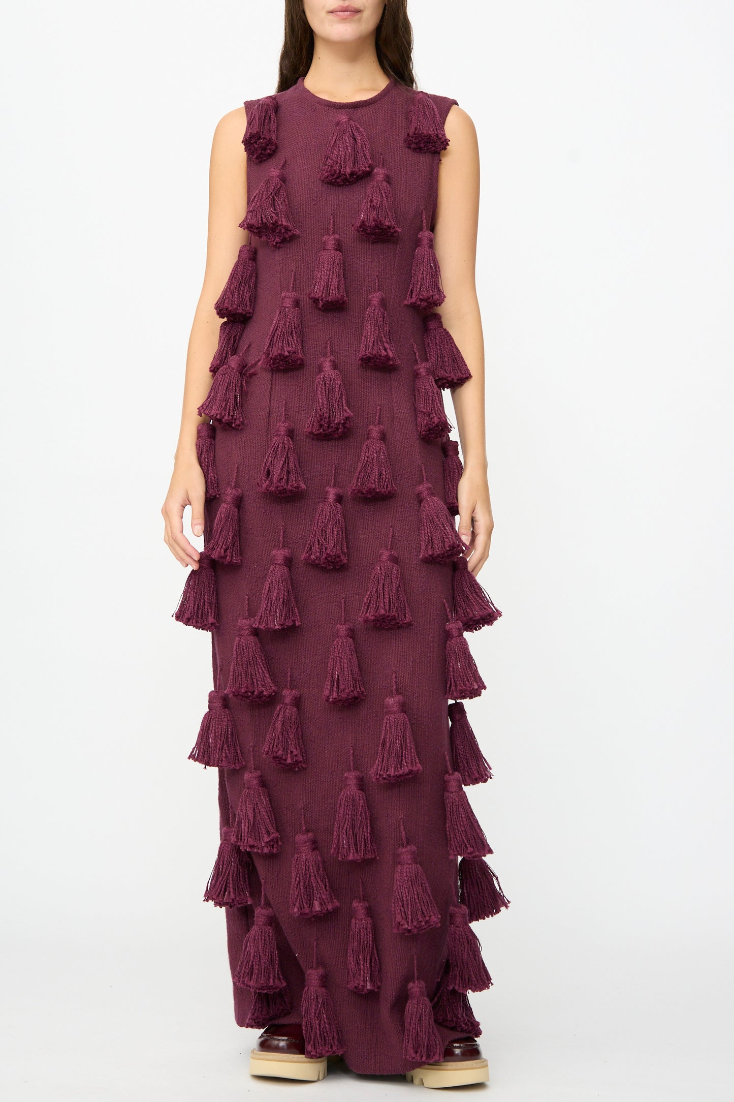 A woman models The Pompom Dress in Merlot Flamme by Marrakshi Life, a sleeveless, floor-length maroon maxi dress adorned with bold, evenly spaced tassels. Crafted from handwoven cotton and handmade in Morocco, she stands against a plain white background.
