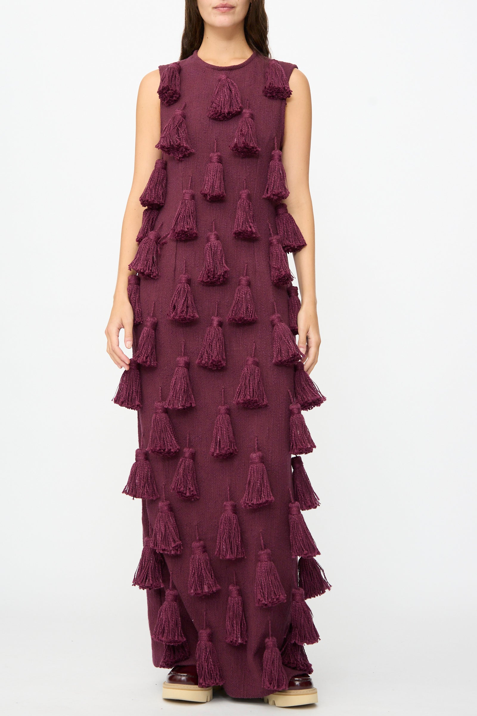 A woman models The Pompom Dress in Merlot Flamme by Marrakshi Life, a sleeveless, floor-length maroon maxi dress adorned with bold, evenly spaced tassels. Crafted from handwoven cotton and handmade in Morocco, she stands against a plain white background.