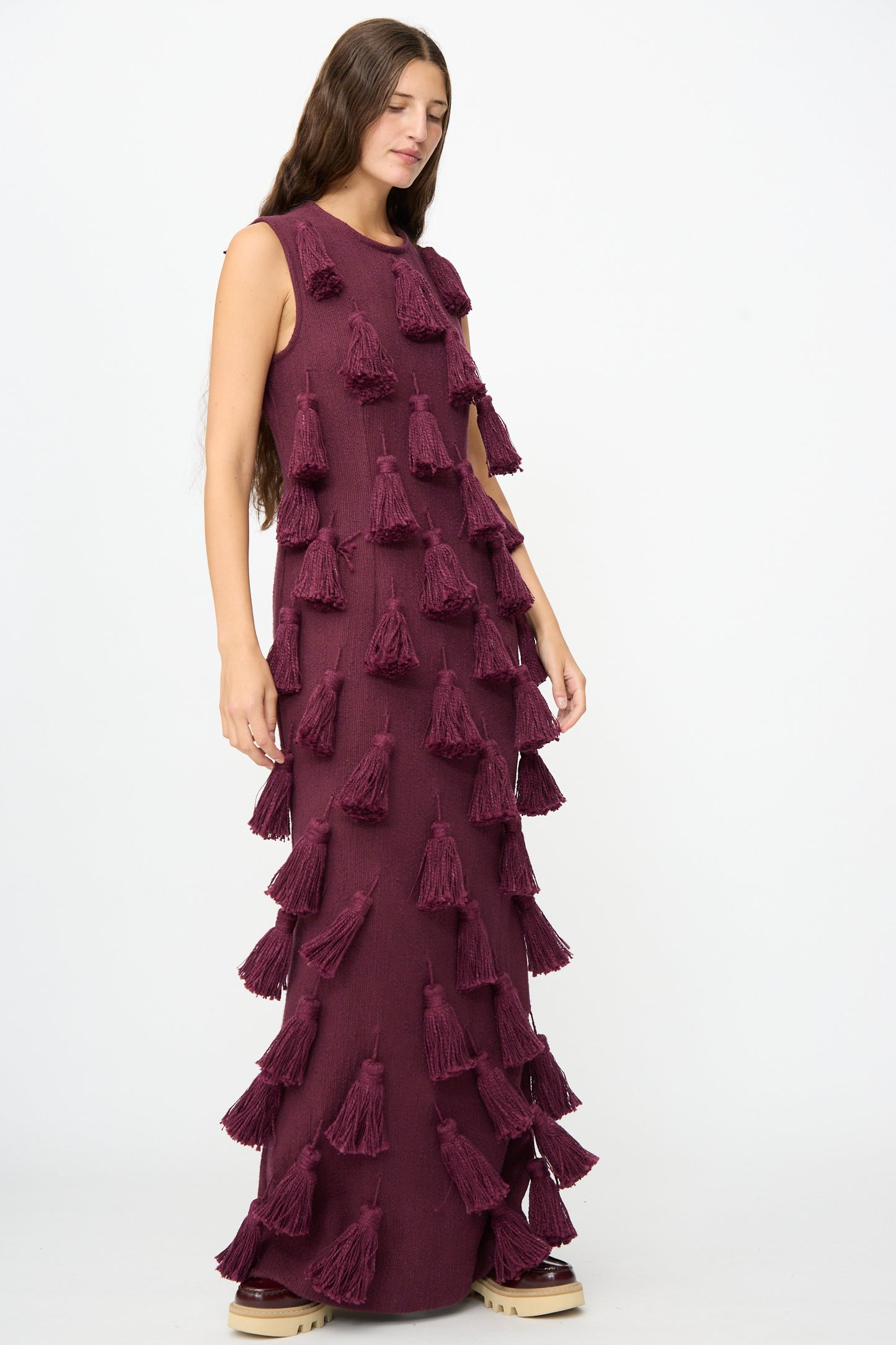 A woman stands against a plain white background wearing Marrakshi Life’s The Pompom Dress in Merlot Flamme—a floor-length, sleeveless maxi dress made from handwoven cotton and embellished with multiple large tassels.