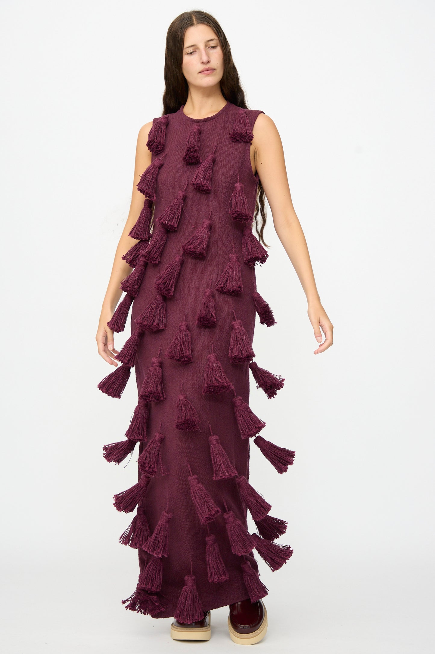 A woman stands with eyes closed against a plain white background, wearing The Pompom Dress in Merlot Flamme by Marrakshi Life—a beautifully handmade, sleeveless maroon maxi dress adorned with large tassels and crafted in Morocco from handwoven cotton.