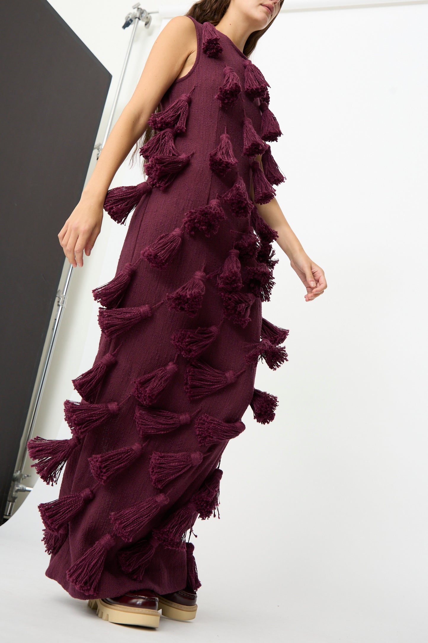 A model wears The Pompom Dress in Merlot Flamme by Marrakshi Life—a floor-length, sleeveless maxi dress in a rich burgundy shade, featuring large, textured tassels and crafted from handwoven cotton in Morocco—standing against a plain studio backdrop.