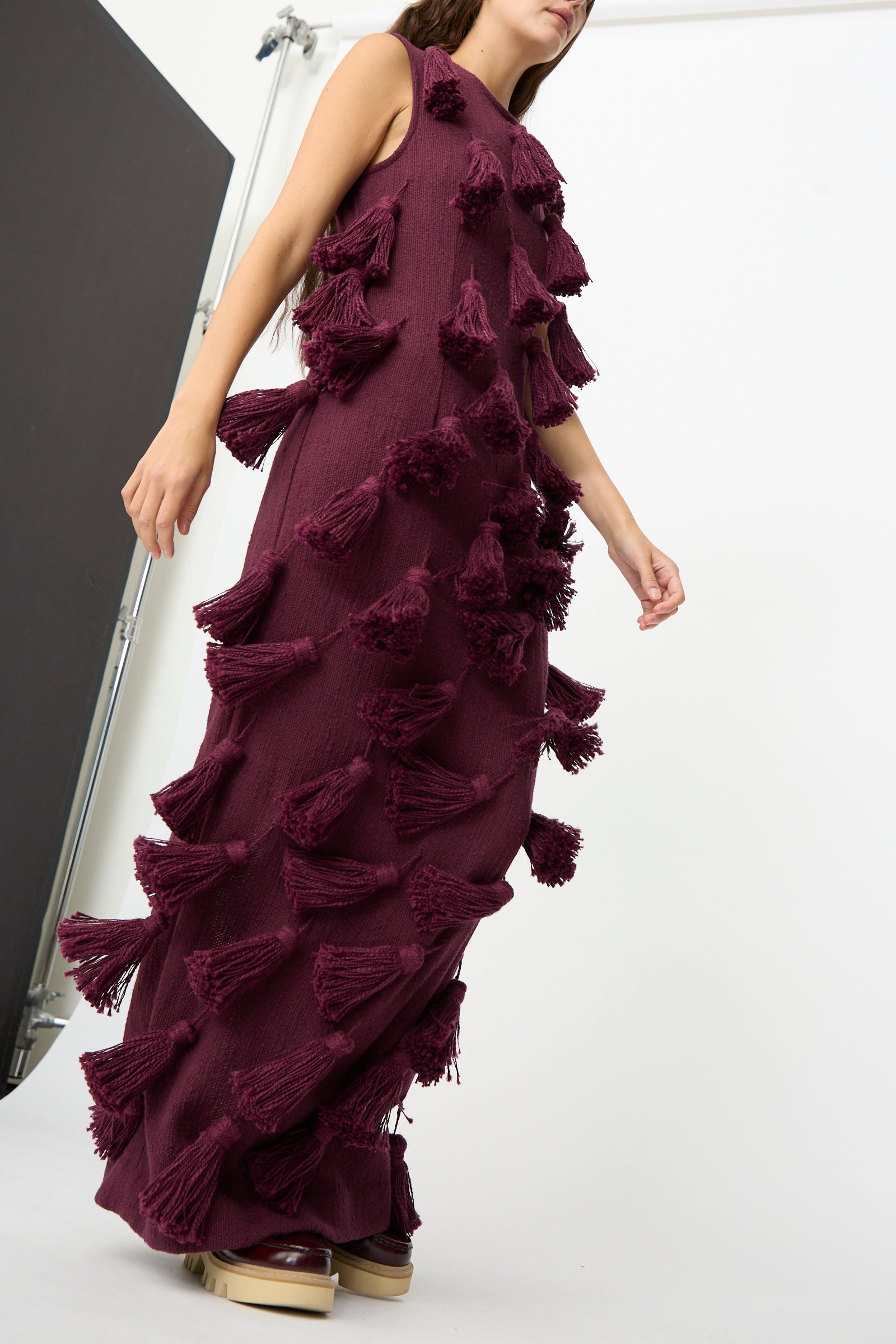 A model wears The Pompom Dress in Merlot Flamme by Marrakshi Life—a floor-length, sleeveless maxi dress in a rich burgundy shade, featuring large, textured tassels and crafted from handwoven cotton in Morocco—standing against a plain studio backdrop.
