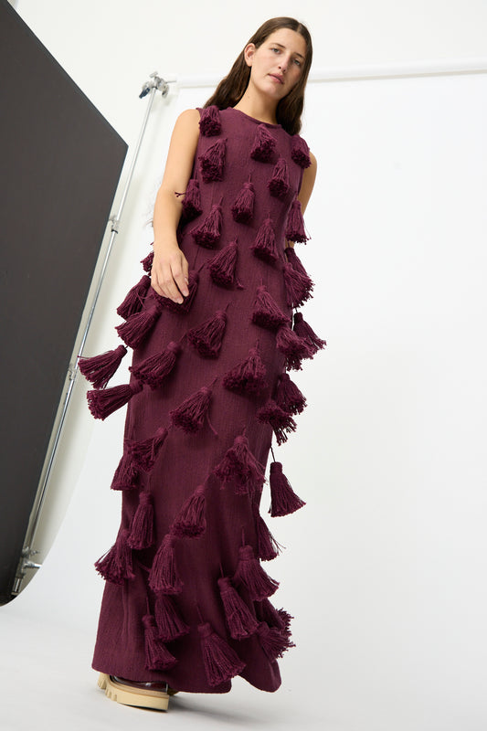A woman stands against a simple studio backdrop, wearing The Pompom Dress in Merlot Flamme by Marrakshi Life—a sleeveless, floor-length maxi dress, handmade in Morocco from handwoven cotton and adorned with large tasseled pom-poms.