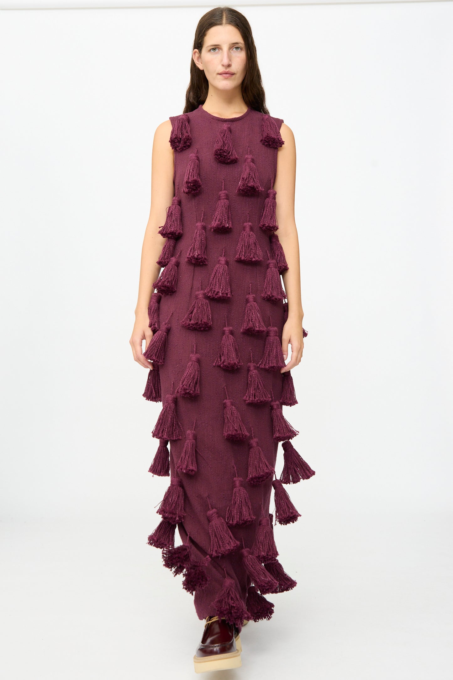 A person stands against a plain white background wearing the Marrakshi Life Pompom Dress in Merlot Flamme — a floor-length, sleeveless maxi dress in rich maroon, crafted from handwoven cotton and adorned with multiple large tassels. This exquisite piece showcases detailed, handmade craftsmanship from Morocco.
