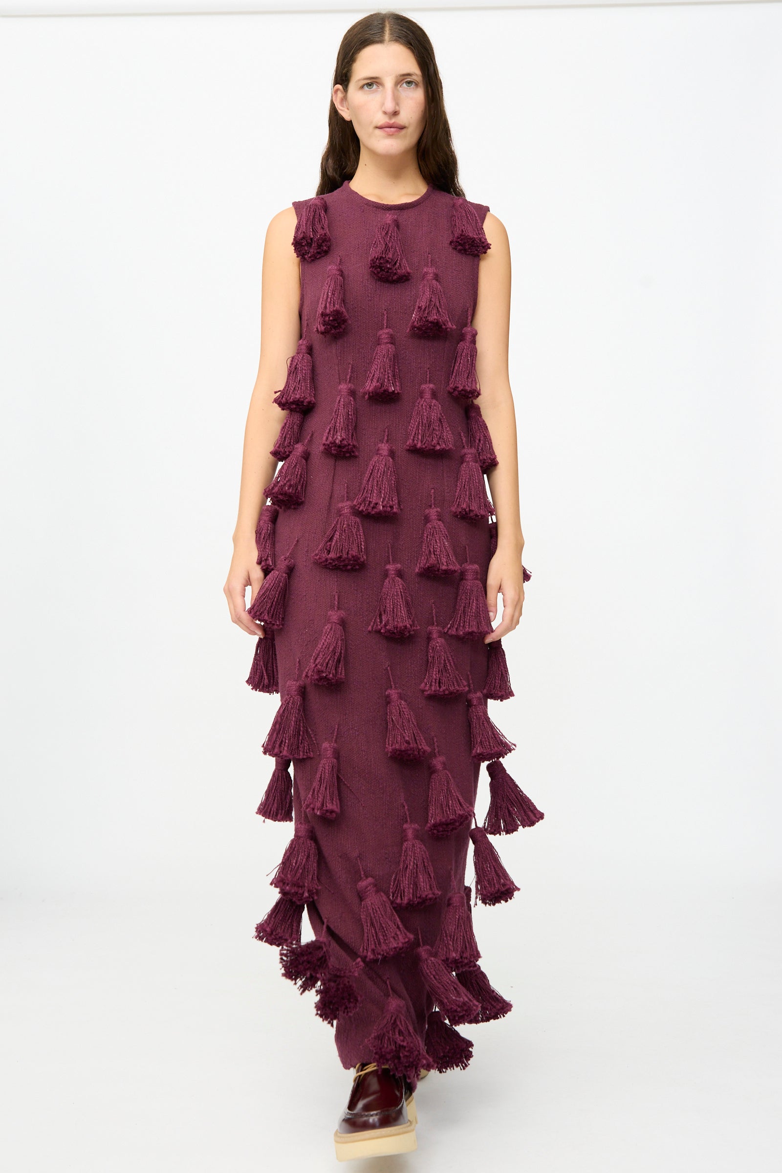 A person stands against a plain white background wearing the Marrakshi Life Pompom Dress in Merlot Flamme — a floor-length, sleeveless maxi dress in rich maroon, crafted from handwoven cotton and adorned with multiple large tassels. This exquisite piece showcases detailed, handmade craftsmanship from Morocco.