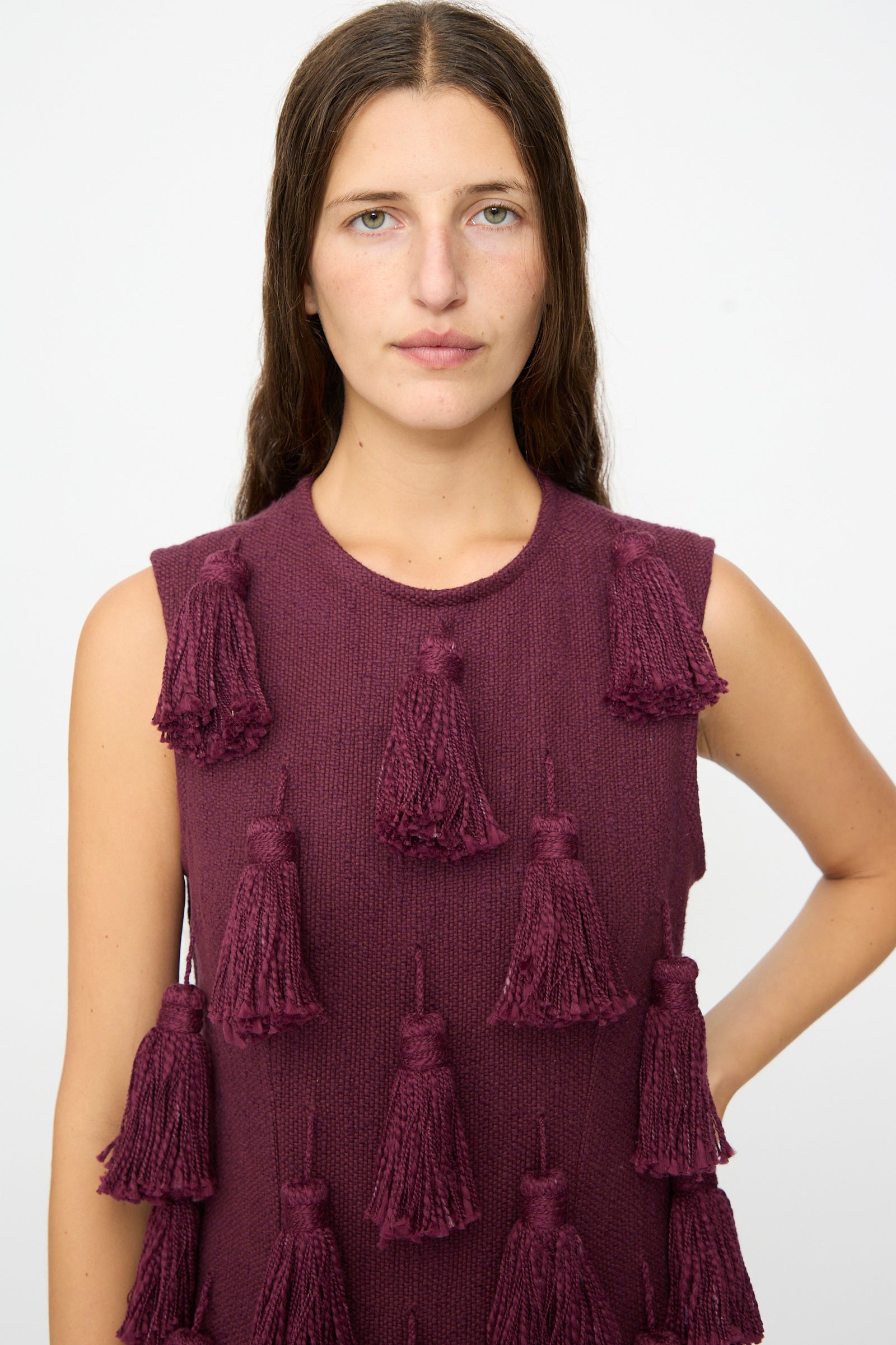 A woman with long brown hair wears The Pompom Dress in Merlot Flamme by Marrakshi Life, a sleeveless, handmade piece from Morocco adorned with large tassels. She stands against a plain white background with one arm bent and a neutral expression.