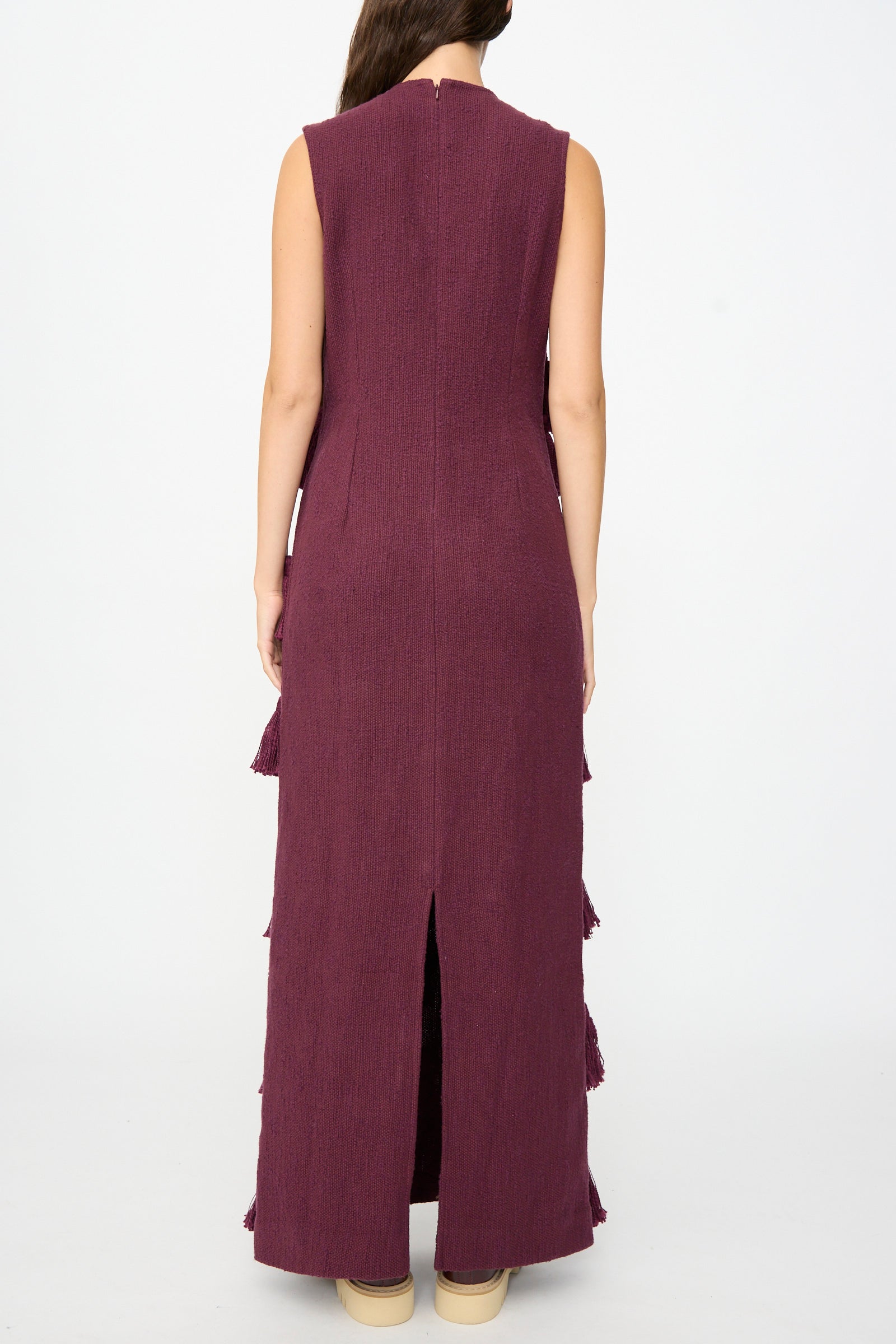 A person stands facing away, wearing Marrakshi Life’s The Pompom Dress in Merlot Flamme—a sleeveless maxi crafted from textured, handwoven cotton in purple with a back slit—paired with beige shoes. This unique piece is handmade in Morocco.