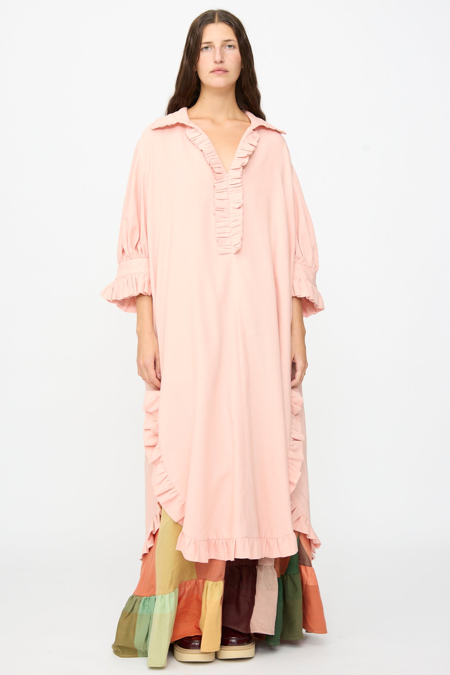 A person stands against a plain background wearing Marrakshi Life’s The Ruffled Tunic in Flesh—an oversized, handwoven cotton tunic in pale pink with ruffled detailing—layered over a tiered skirt showcasing blocks of green, tan, red, and brown, finished with cream platform shoes. The ensemble’s relaxed silhouette underscores its artisanal craftsmanship and is handmade in Morocco.