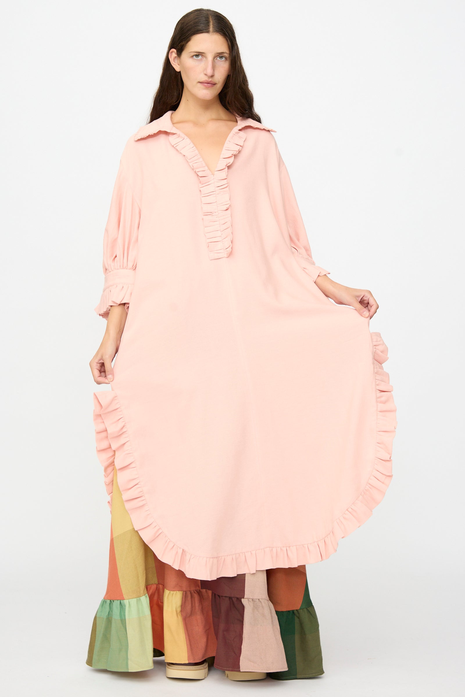 A woman wearing The Ruffled Tunic in Flesh by Marrakshi Life, a loose, handwoven cotton piece in light pink with ruffled edges, layered over a color-blocked tiered skirt. She stands against a plain white background, highlighting the oversized fit and artisan craftsmanship that make this handmade-in-Morocco tunic uniquely charming.
