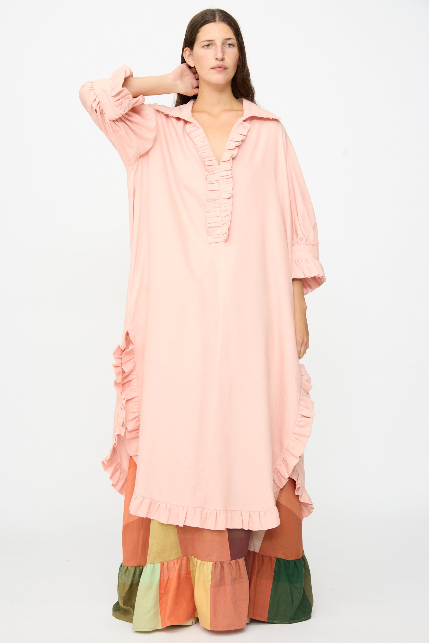 A model stands against a plain white background, wearing Marrakshi Life’s The Ruffled Tunic in Flesh—a loose, long pink dress with ruffled edges—styled in an oversized fit over a tiered, multicolored skirt.
