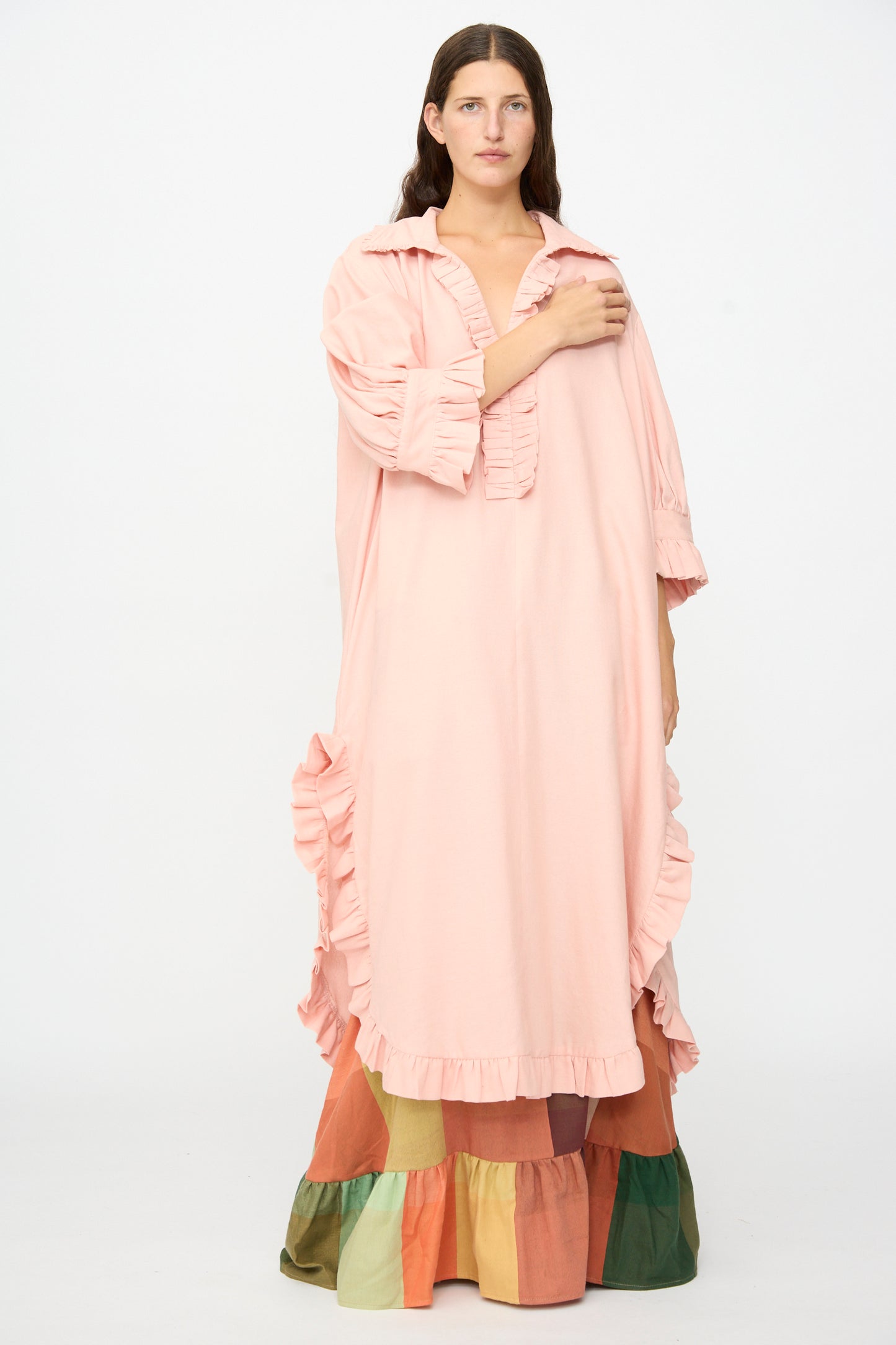 A person stands against a plain white background wearing The Ruffled Tunic in Flesh by Marrakshi Life, an oversized, light pink dress with ruffled details, layered over a colorful tiered skirt and reminiscent of a handwoven cotton tunic handmade in Morocco.