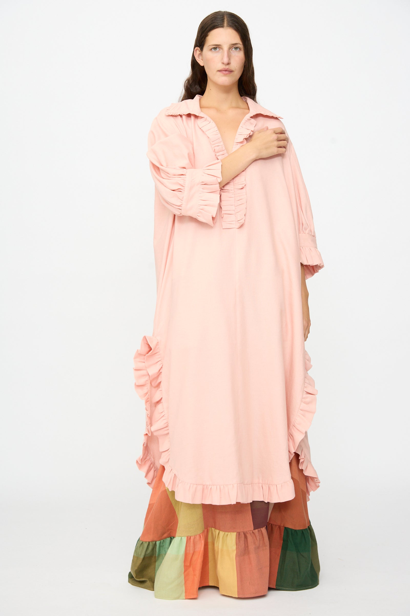 A person stands against a plain white background wearing The Ruffled Tunic in Flesh by Marrakshi Life, an oversized, light pink dress with ruffled details, layered over a colorful tiered skirt and reminiscent of a handwoven cotton tunic handmade in Morocco.