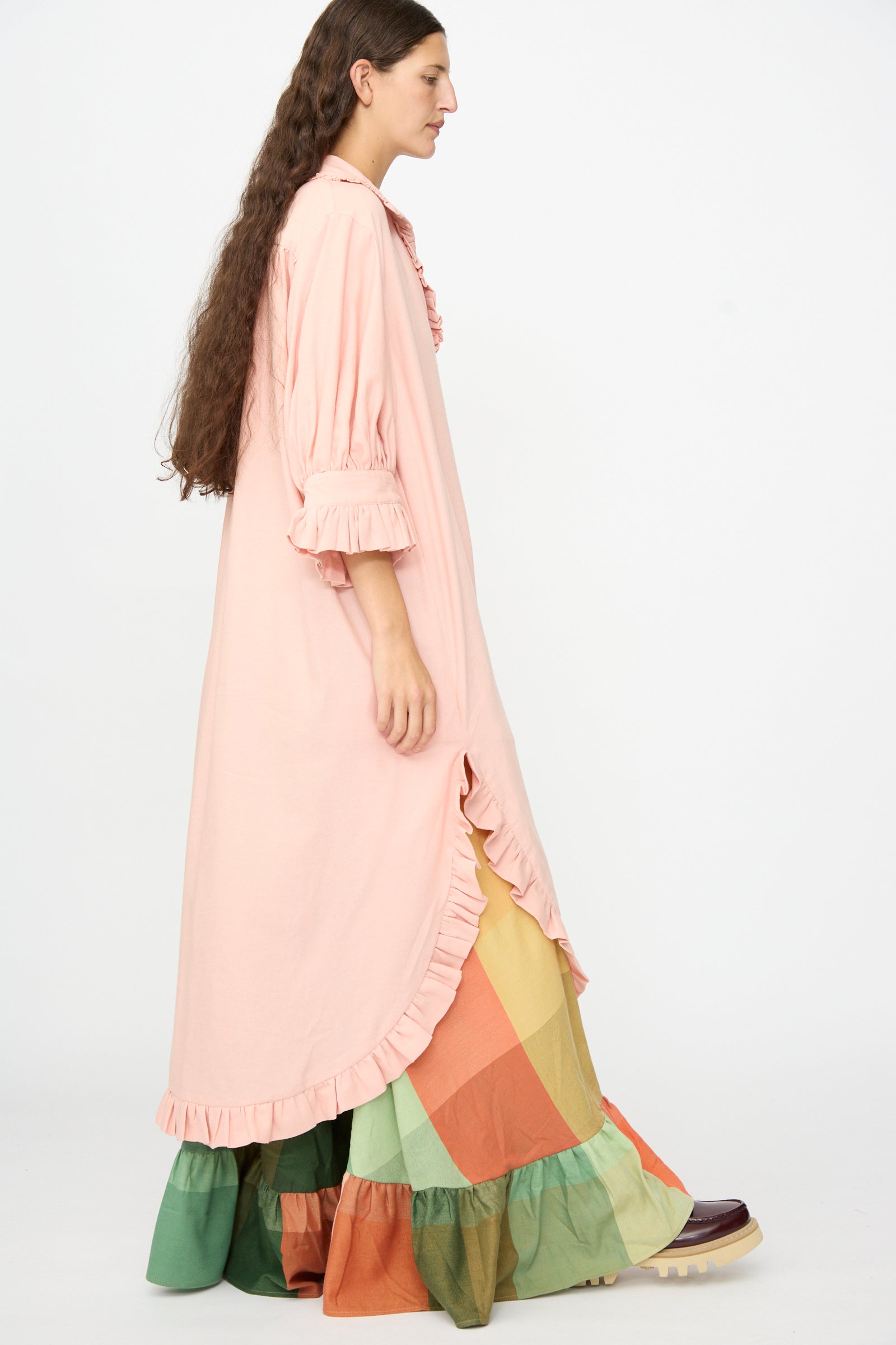 A person wears The Ruffled Tunic in Flesh by Marrakshi Life—an oversized, long pink ruffled dress featuring a checker-patterned skirt in green, orange, and yellow tones—paired with brown shoes and set against a plain white background. The ensemble captures the relaxed appeal of a handwoven cotton tunic.