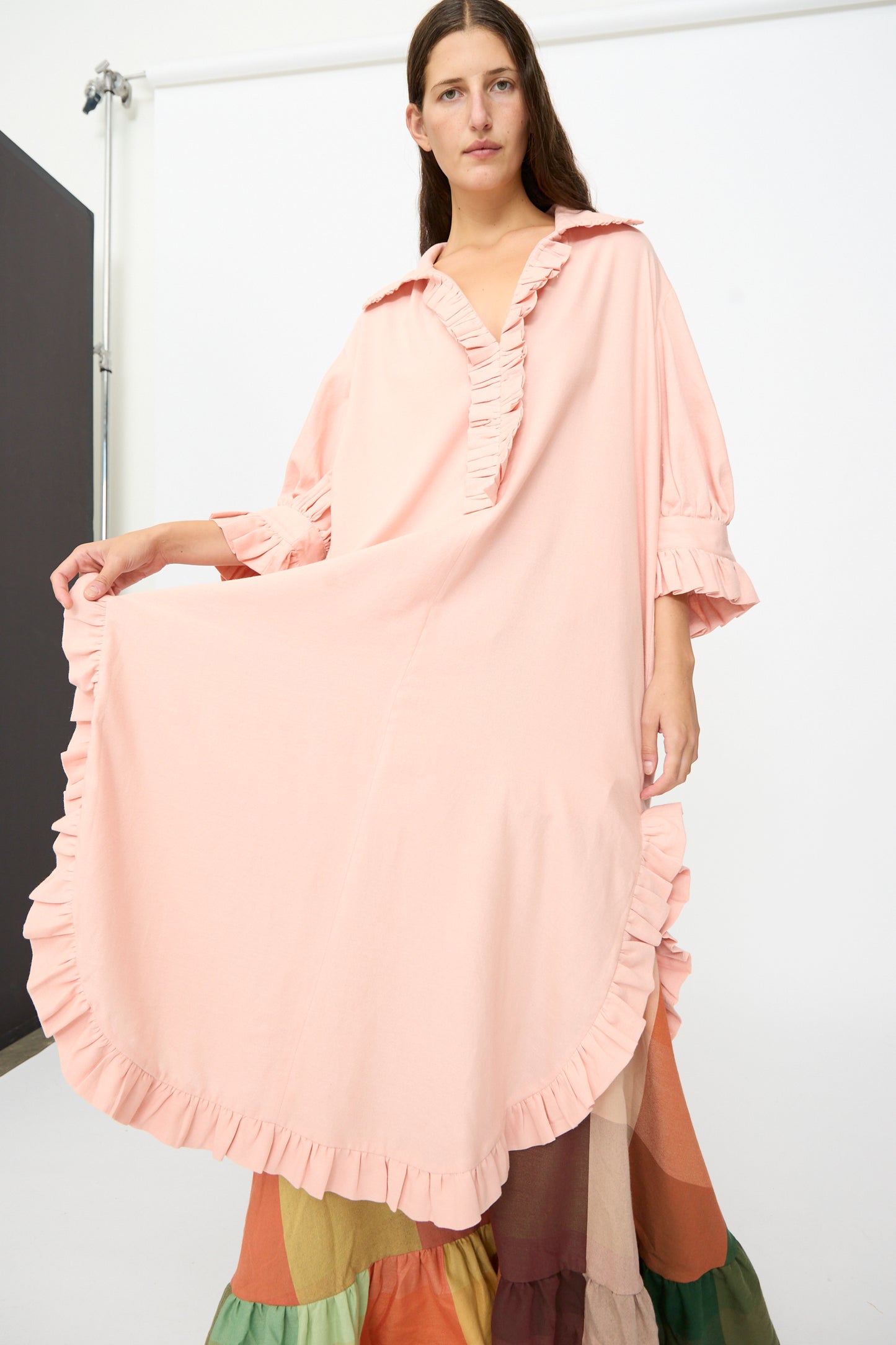 A person stands against a plain background wearing The Ruffled Tunic in Flesh by Marrakshi Life—a loose, handwoven cotton piece featuring ruffled edges and a multi-colored skirt, designed with an oversized fit and handmade in Morocco.