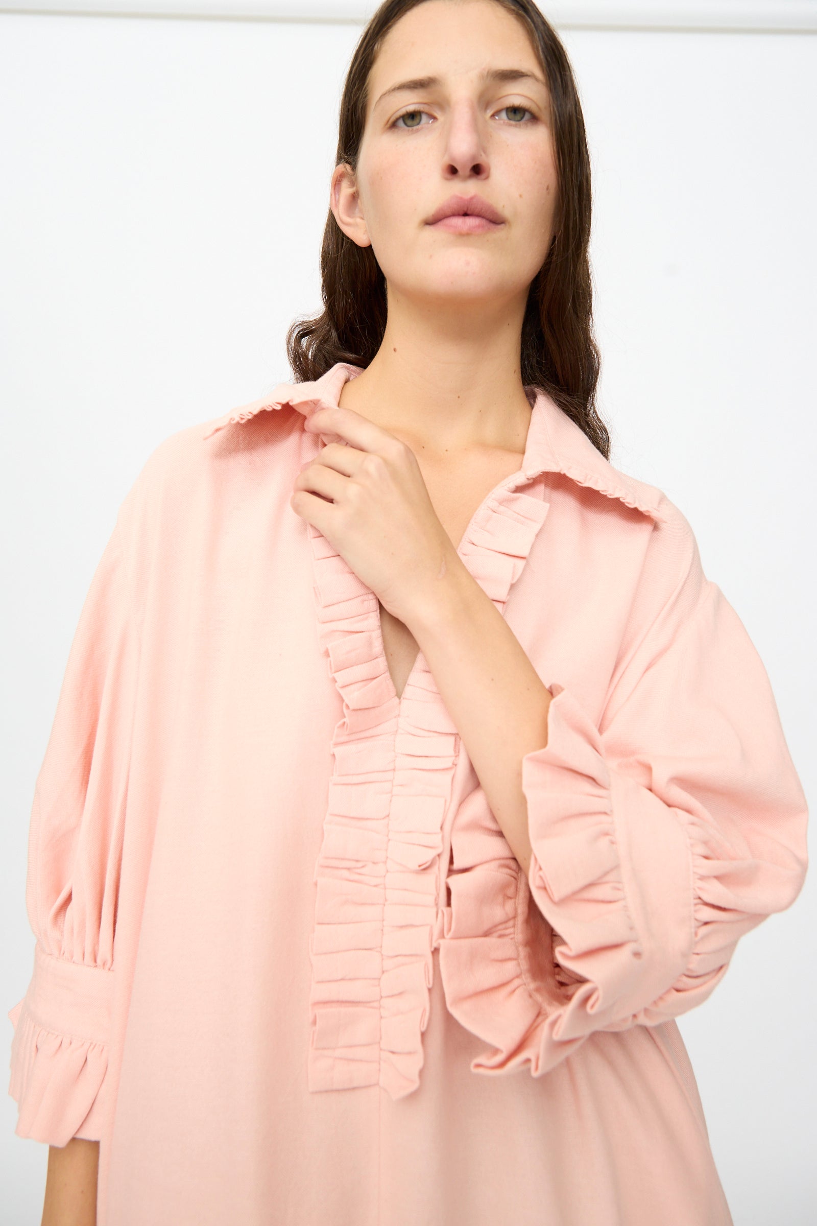 A person wearing Marrakshi Life’s The Ruffled Tunic in Flesh—a loose, handwoven cotton tunic in light pink with ruffled details on the collar and sleeves—stands against a plain white background.