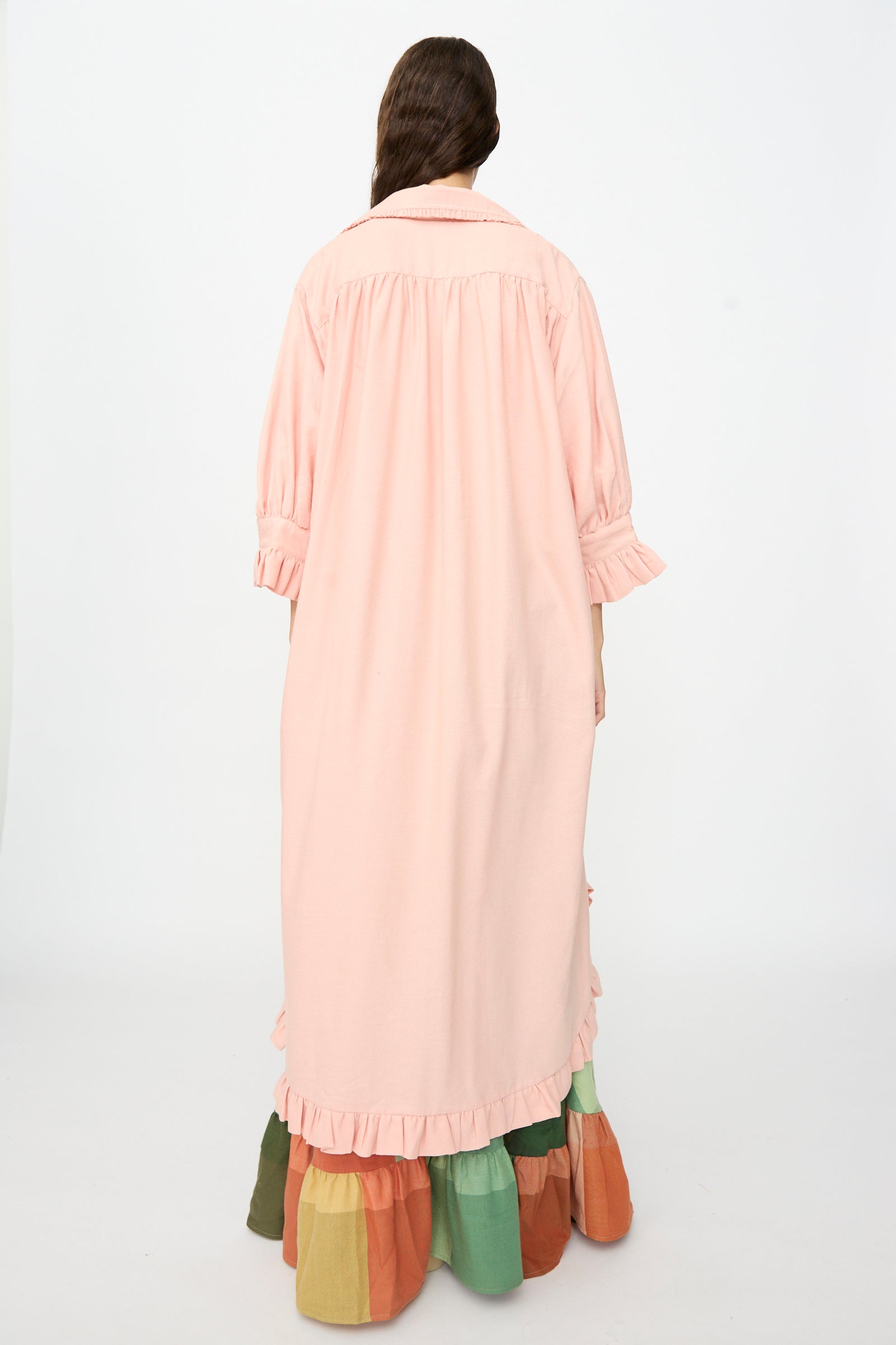 A person with long brown hair stands facing away, wearing Marrakshi Life’s The Ruffled Tunic in Flesh—an oversized, handwoven cotton dress handmade in Morocco, featuring ruffled cuffs and hem. The tunic is styled over a multicolored tiered skirt against a plain white background.