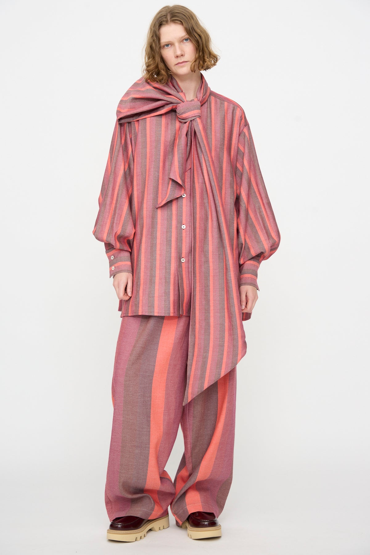 A person wearing Marrakshi Life’s The Scarf Blouse in Stripe Multi—an oversized, pink and orange striped cotton top with a large matching bow—paired with wide-legged pants and platform shoes, stands against a plain white background.