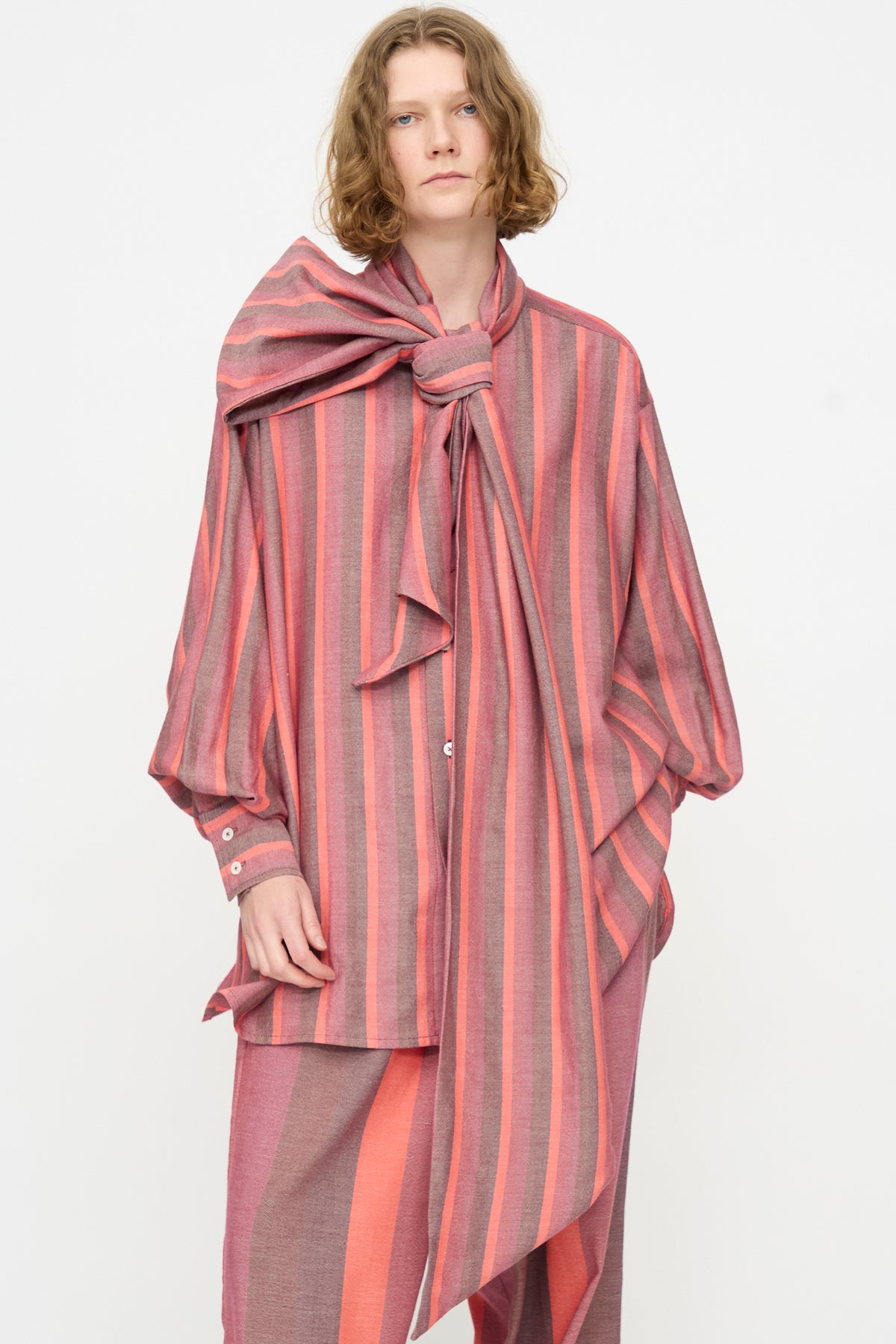 A person stands against a plain white background, wearing Marrakshi Life’s The Scarf Blouse in Stripe Multi—a loose, long-sleeved cotton top in pink and gray stripes featuring an oversized fit and a statement bow detail at the neck.