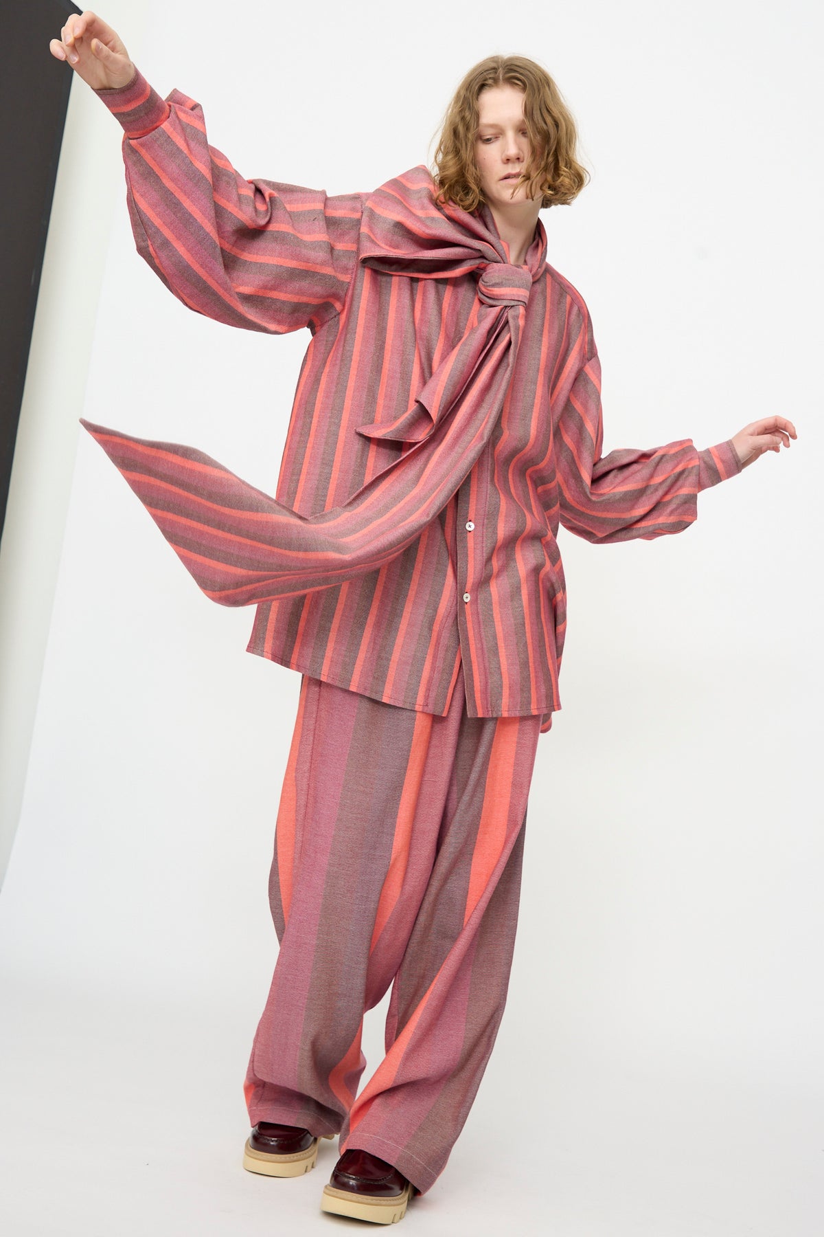 Wearing Marrakshi Life’s The Scarf Blouse in Stripe Multi—a loose cotton blouse with pink and orange stripes, oversized fit, and matching pants—this person is accented by a large scarf bow and stands against a plain white background.