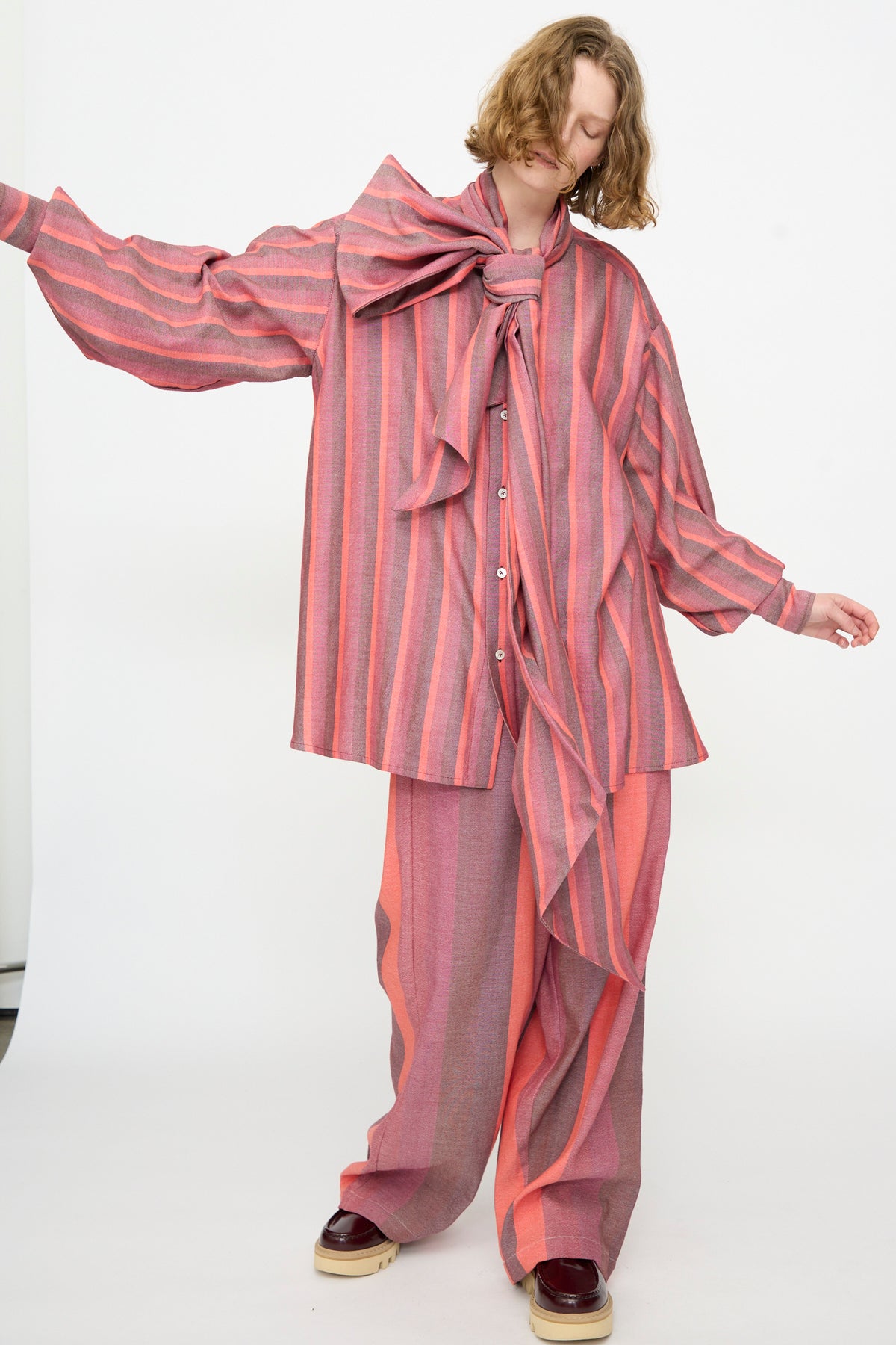 A person wearing The Scarf Blouse in Stripe Multi by Marrakshi Life—a loose, oversized cotton blouse with pink and gray stripes, long sleeves, matching pants, and a large bow at the collar—stands against a plain white background.