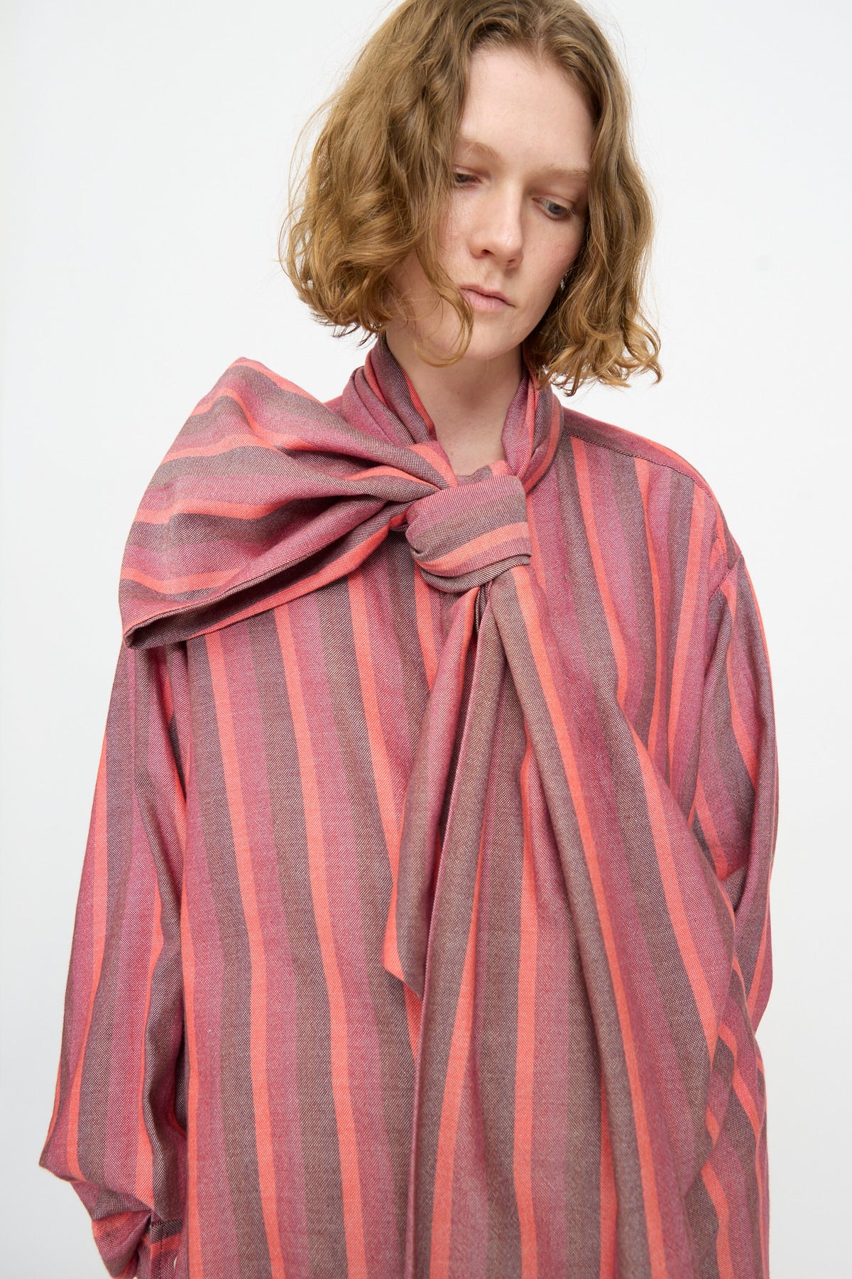 A person with short, wavy hair is seen standing against a plain white background, wearing Marrakshi Life’s The Scarf Blouse in Stripe Multi—a loose-fitting pink and gray striped cotton blouse featuring long sleeves and an oversized bow at the neck.