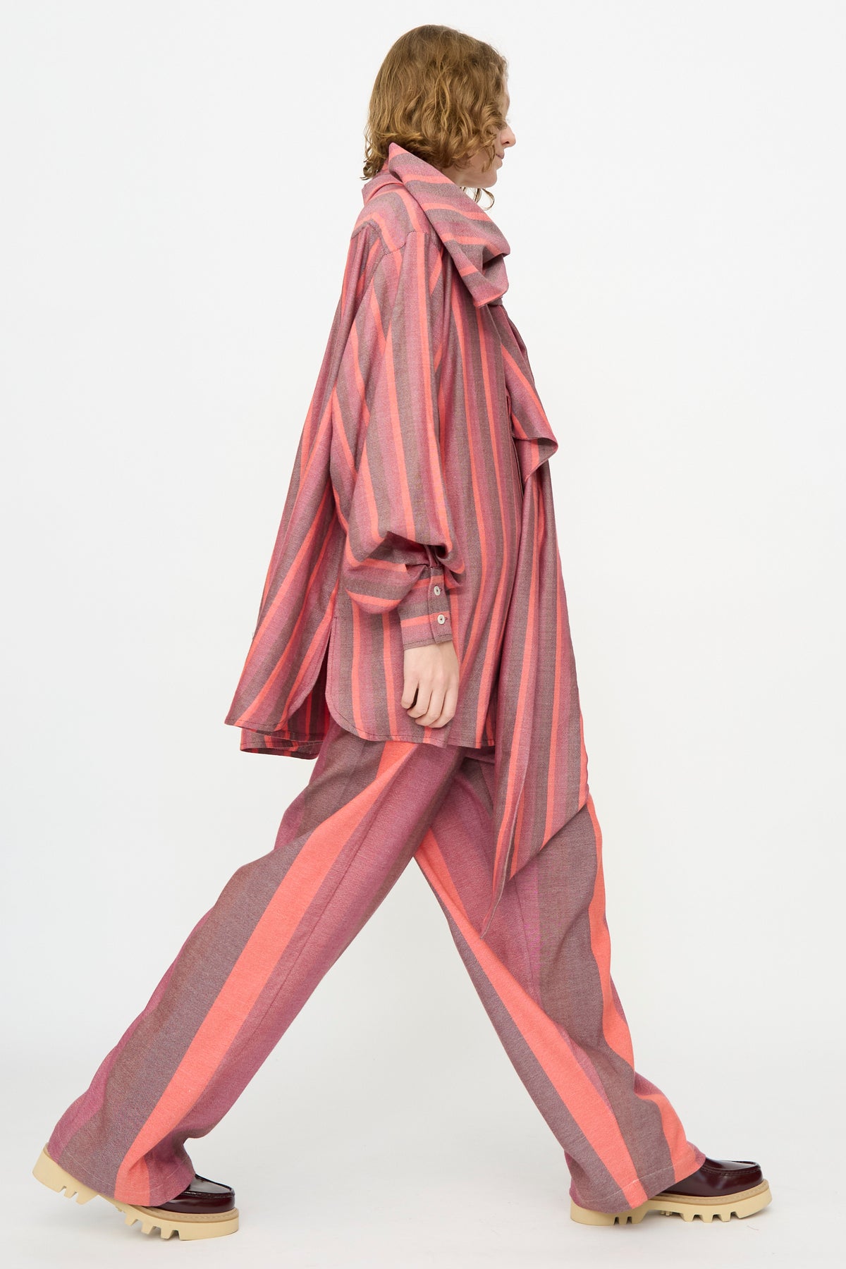 A person in profile wears Marrakshi Life’s The Scarf Blouse in Stripe Multi—an oversized fit, pink and gray striped cotton blouse paired with matching pants and beige-soled burgundy shoes—walking against a plain white background.