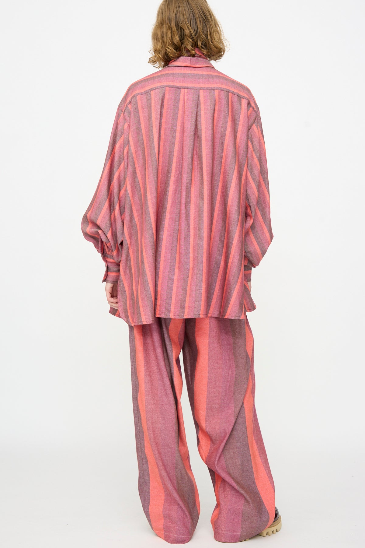 A person with shoulder-length, light brown hair stands with their back to the camera, wearing Marrakshi Life’s The Scarf Blouse in Stripe Multi—an oversized fit striped cotton blouse—paired with matching wide-leg trousers in vertical red and grey stripes.
