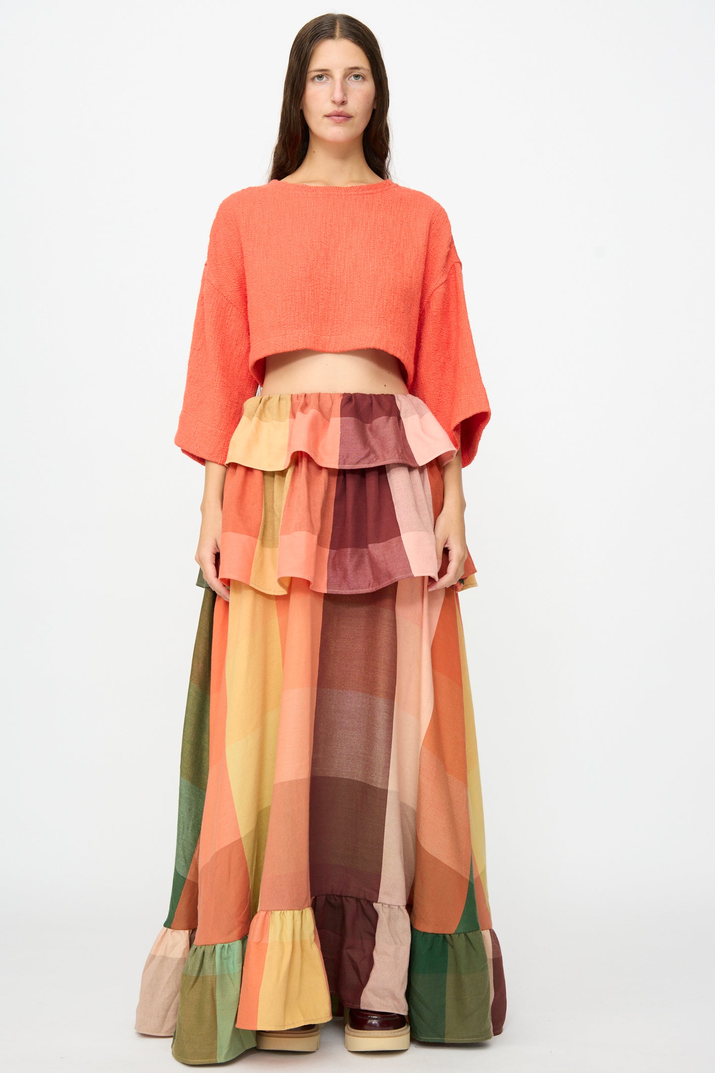 A woman stands facing forward against a plain white background, wearing Marrakshi Life’s The Tiered Ruffle Skirt in Plaid—a long, multicolored patchwork maxi skirt with ruffle details, handmade in Morocco—and an orange cropped top.