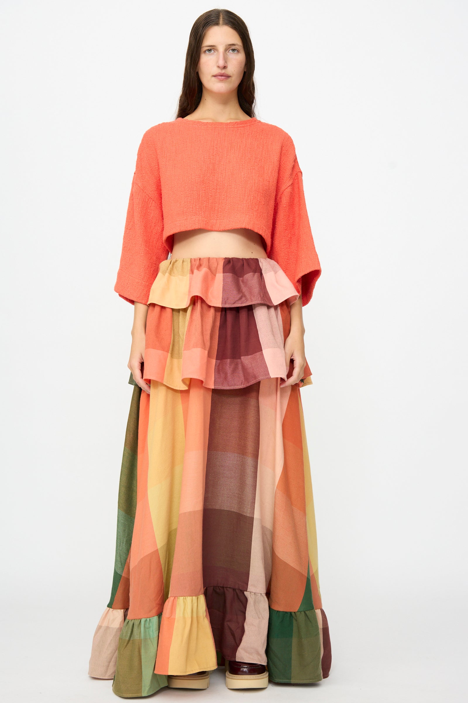 A woman stands facing forward against a plain white background, wearing Marrakshi Life’s The Tiered Ruffle Skirt in Plaid—a long, multicolored patchwork maxi skirt with ruffle details, handmade in Morocco—and an orange cropped top.